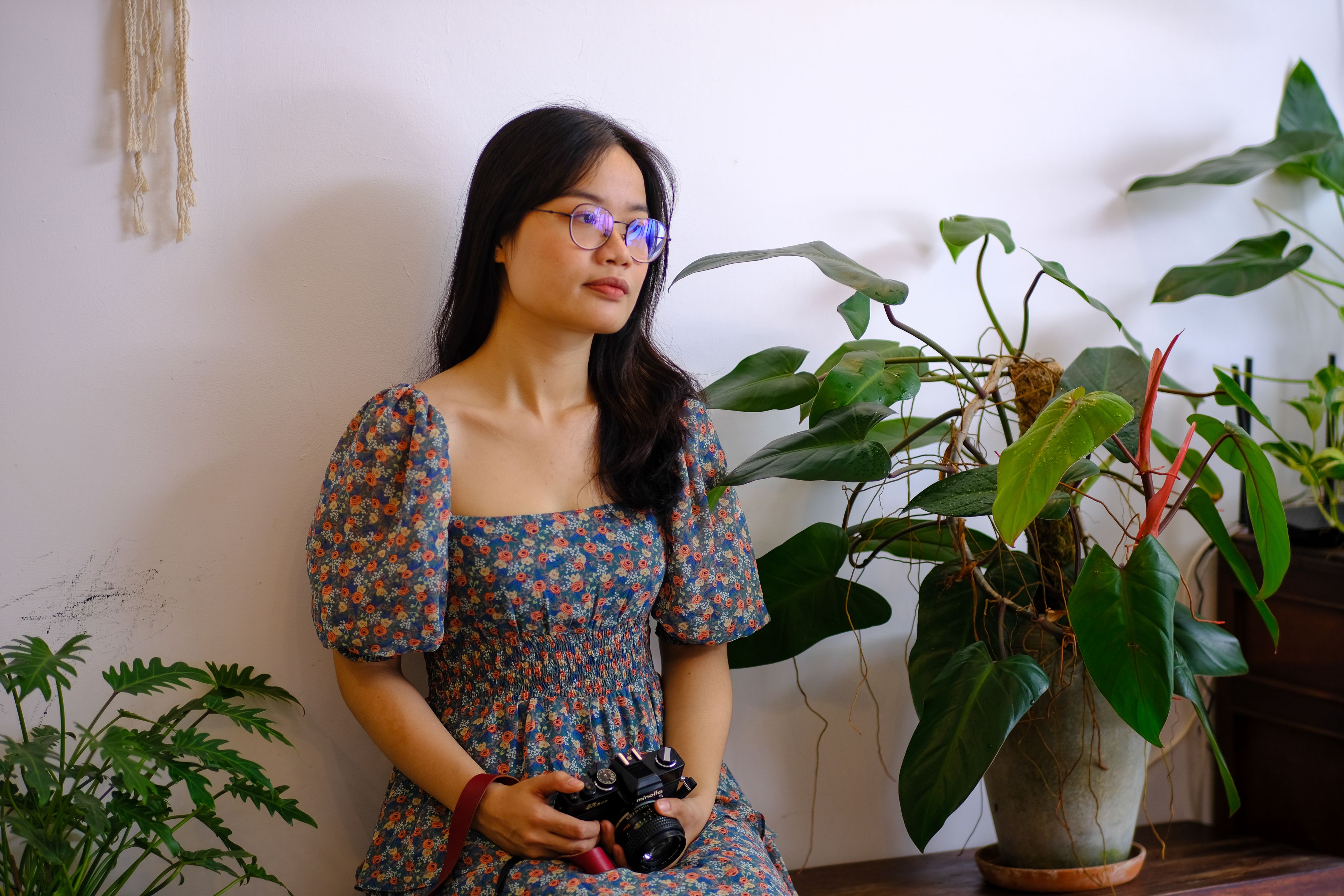 A woman in glasses and floral dress reflects indoors, holding a vintage camera among houseplants.