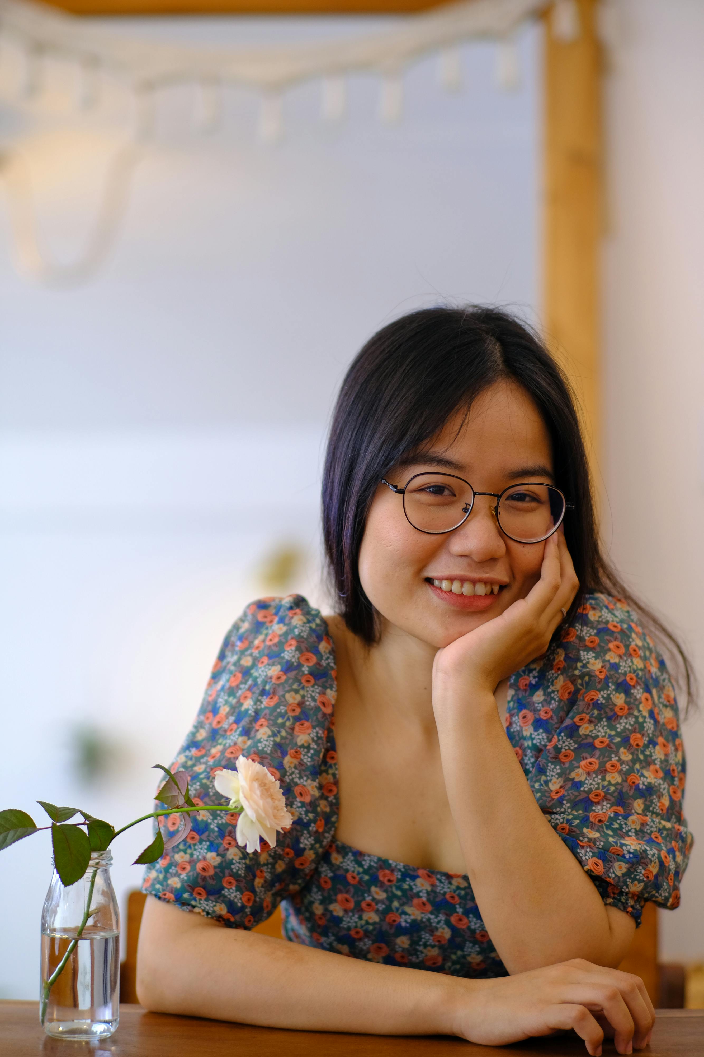 A smiling woman with glasses in a floral dress, captured in a cozy indoor setting in Hue, Vietnam.