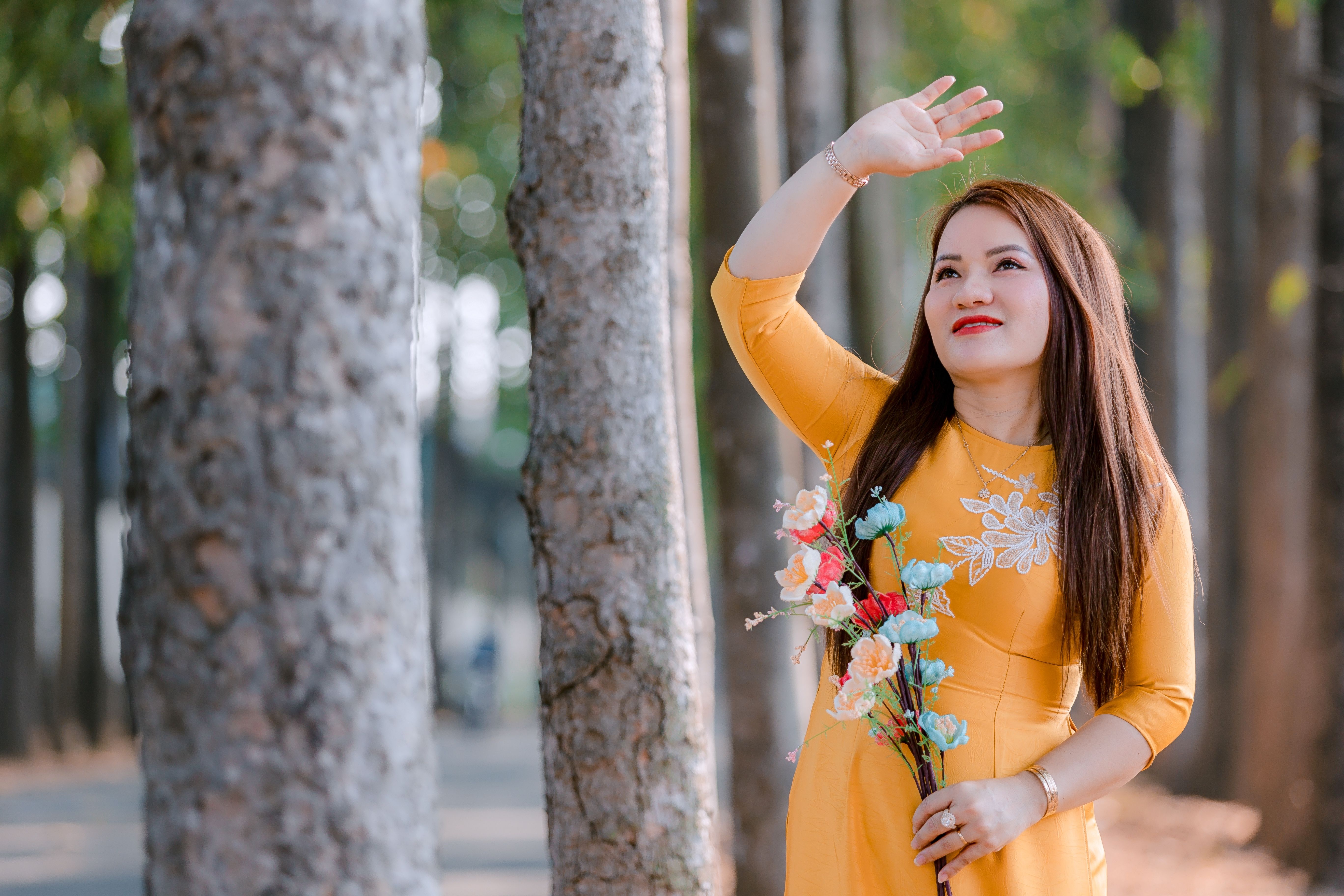 A woman in a yellow dress holding flowers looks up in a peaceful forest setting.