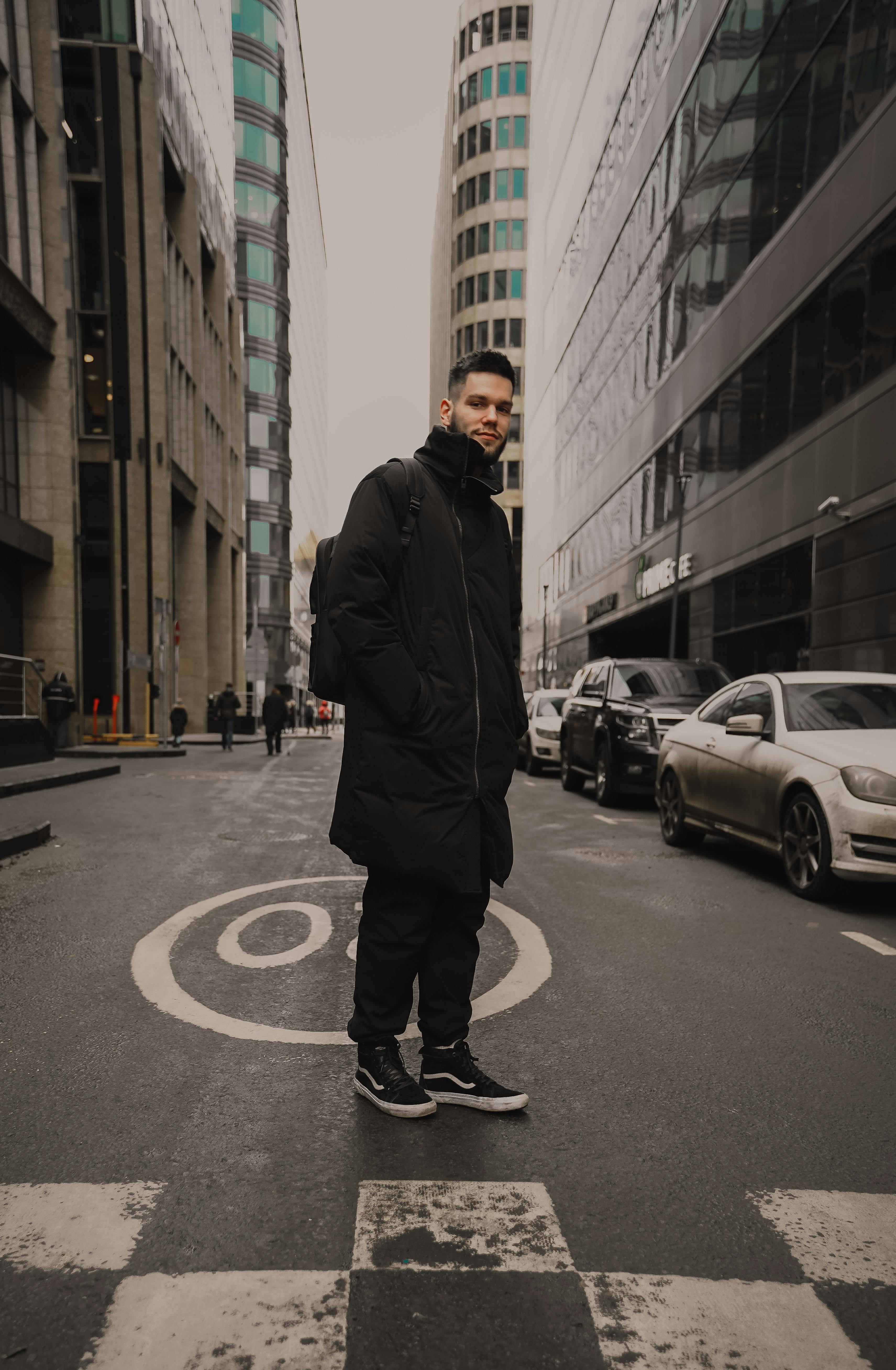 一位时尚男子穿着黑色外套，自信地站在城市街道上，四周环绕着高大的现代建筑