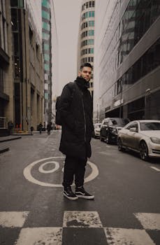 一位时尚男子穿着黑色外套，自信地站在城市街道上，四周环绕着高大的现代建筑