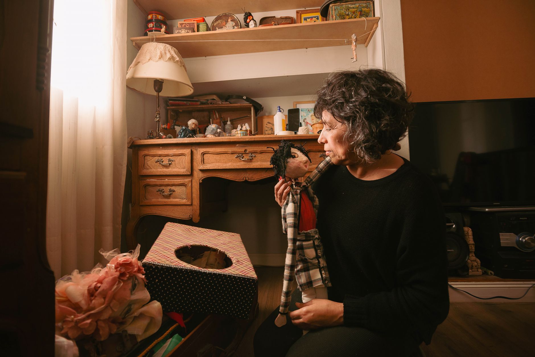 A woman in a softly lit room holds a handmade puppet, surrounded by vintage decor and soft lighting.
