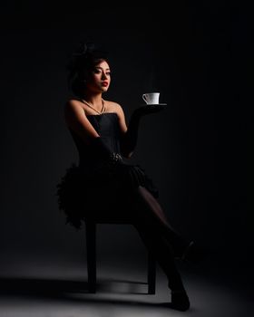 Elegant woman in a vintage black dress holding a steaming teacup in a dramatic pose.