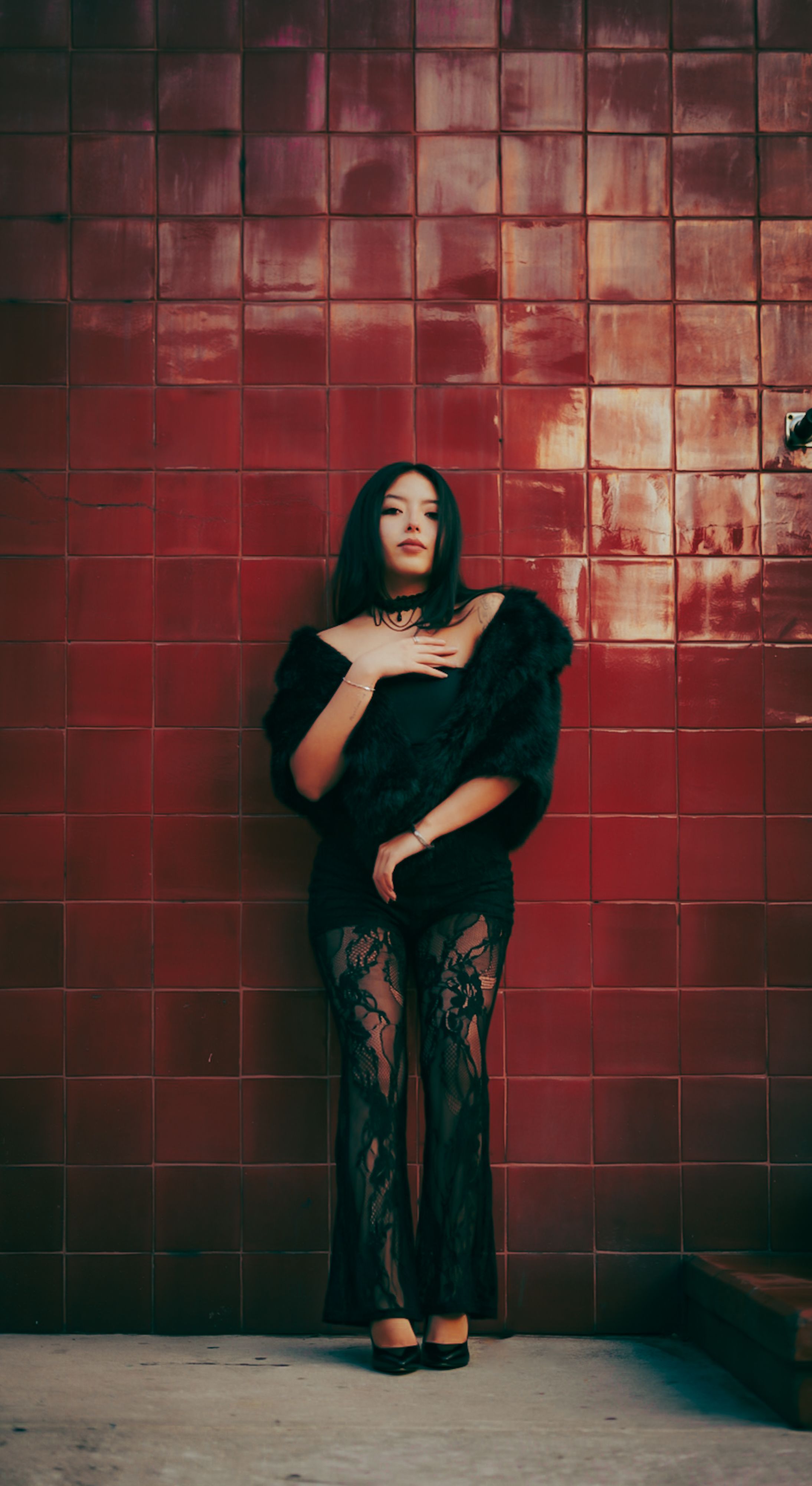Elegant woman in black lace and fur stands against a red tiled wall, exuding confidence and style.