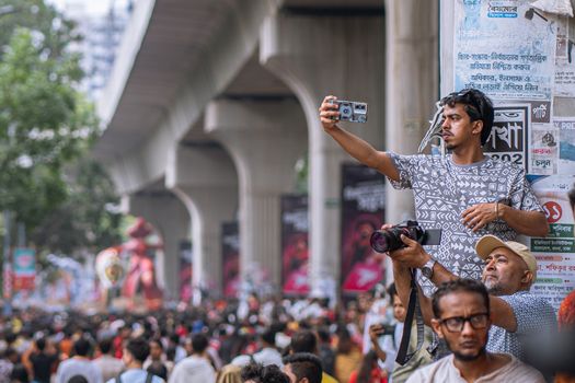Photo by Julian Fernandez A lively street festival with a crowd capturing moments on camera amid colorful decorations and urban infrastructure.