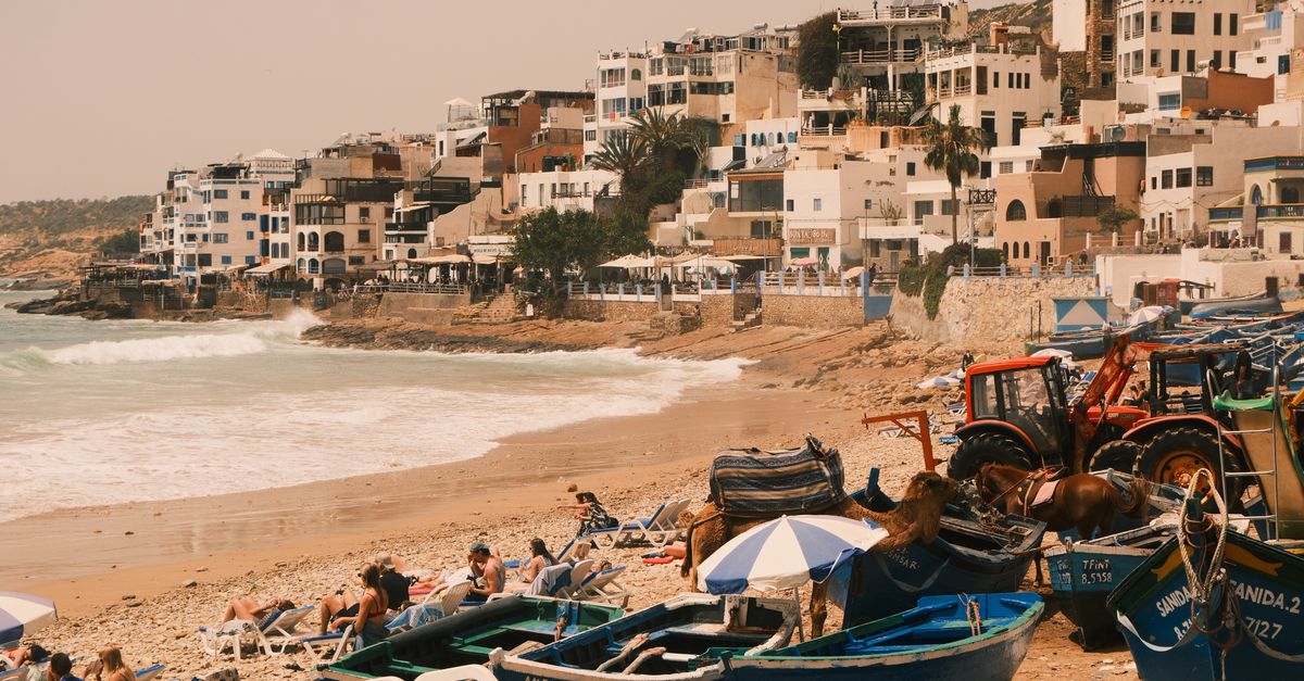 Scenic view of Taghazout beach with colorful boats, sunbathers, and adjacent hillside buildings.