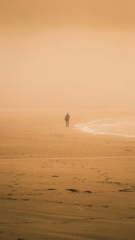 A lone figure walks on a foggy beach during sunrise, creating a serene and introspective scene.