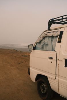 Off-road white van parked on a sandy coastal road with foggy background, suggesting travel adventure.