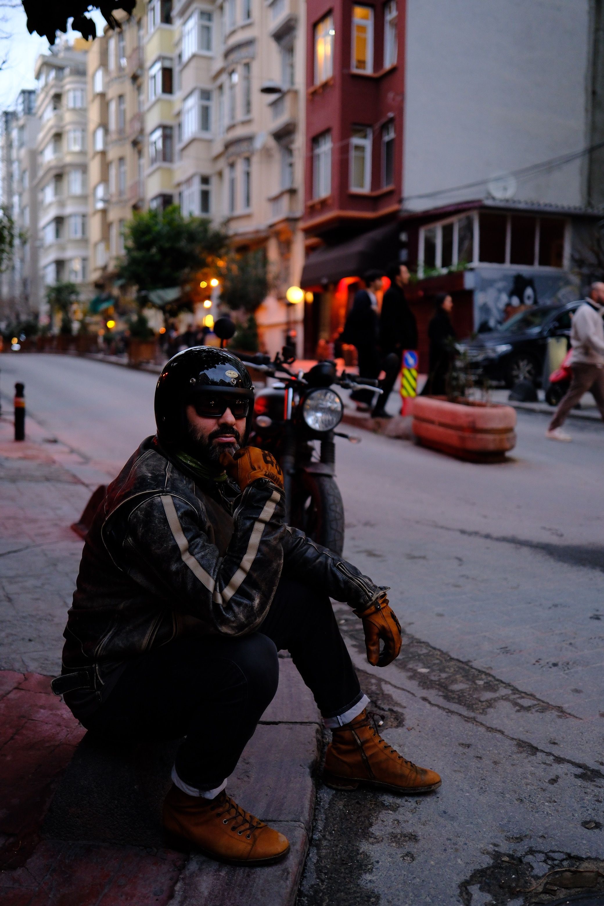 A motorcyclist in gear sits on the sidewalk of a city street at twilight, exuding a moody and cool vibe.