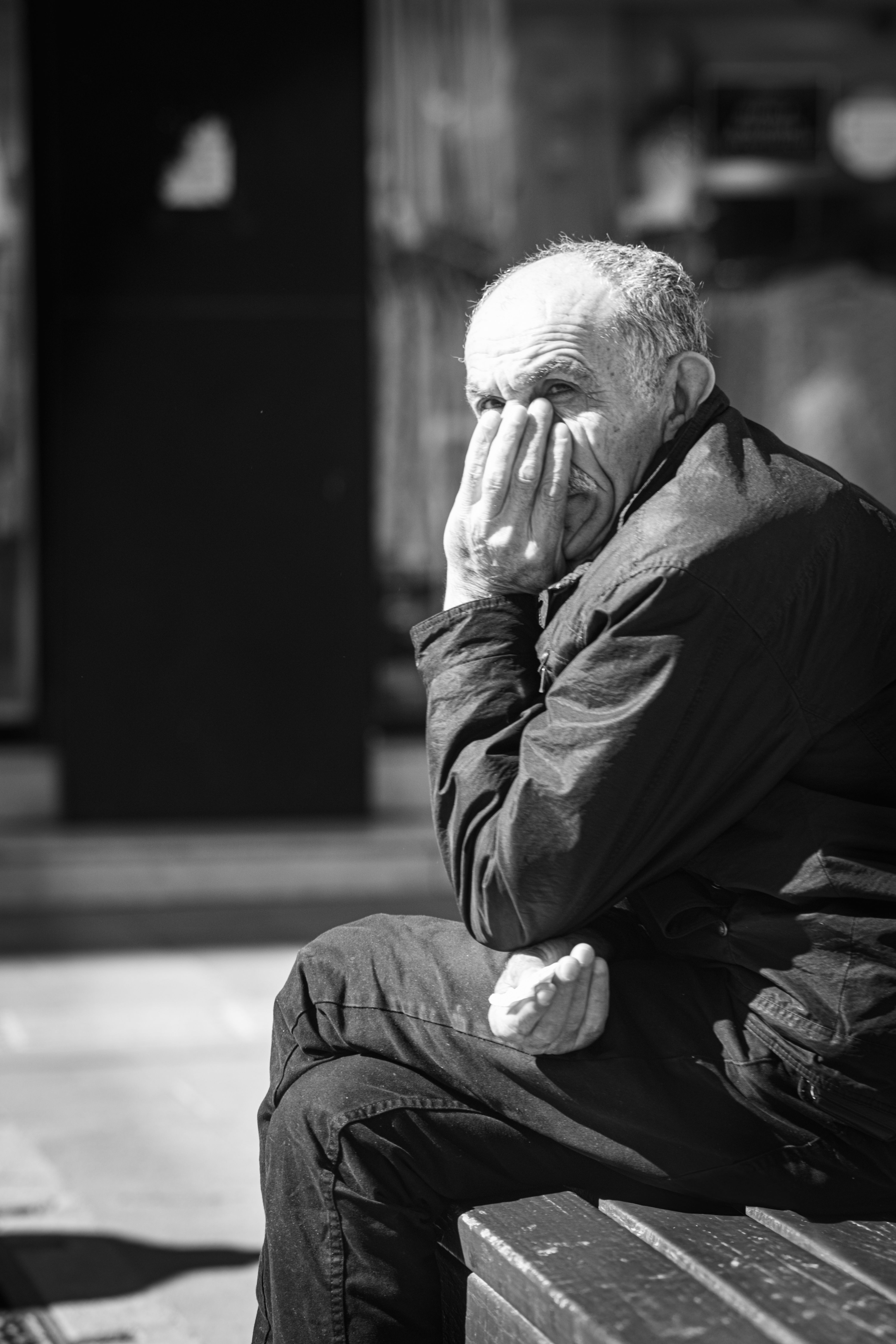 A thoughtful elderly man sits on a bench outdoors, hand on face.