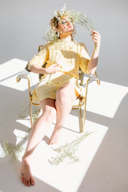 A woman in a yellow dress on a chair, adorned with baby's breath flowers in bright, natural lighting.