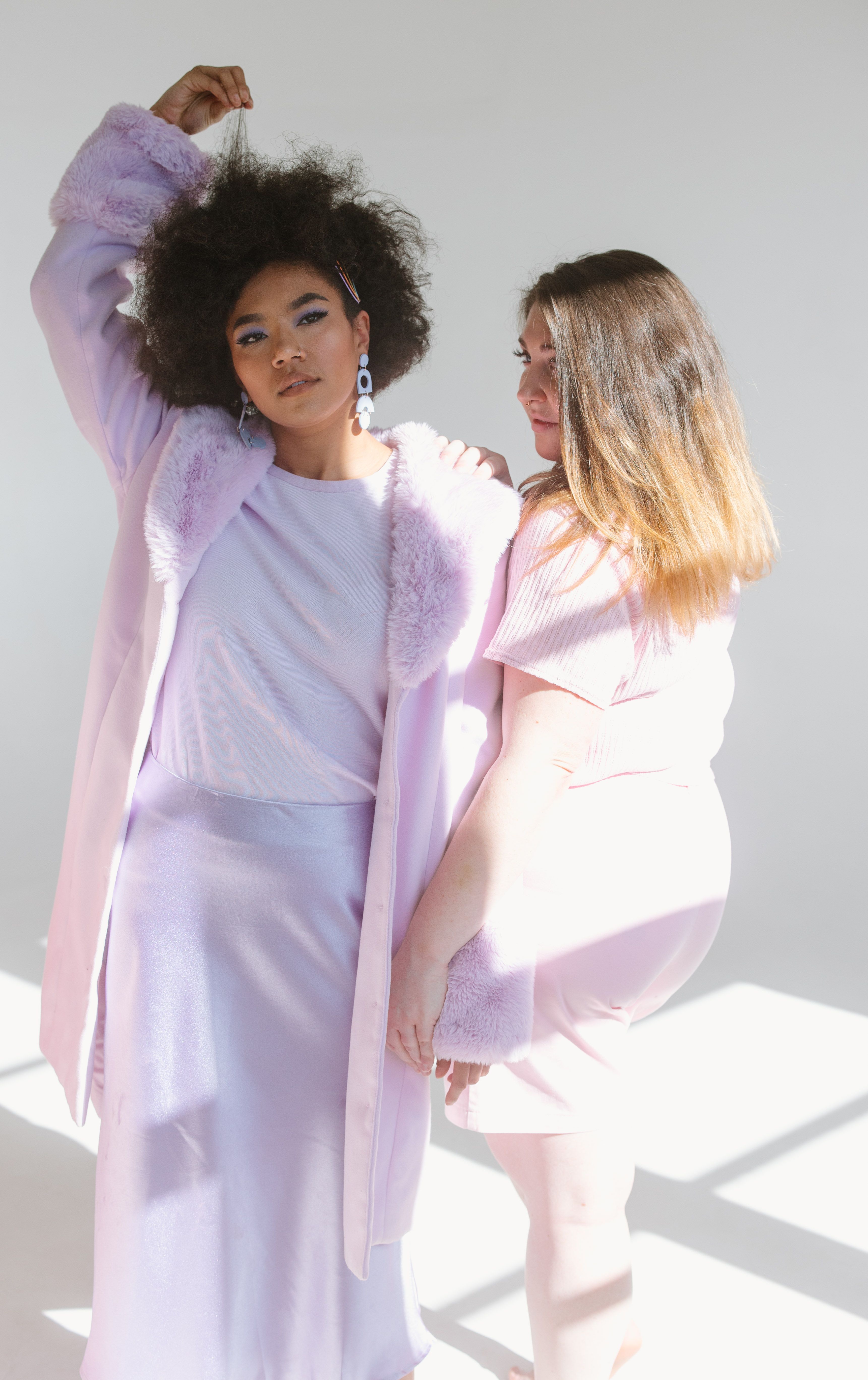 Two stylish women in pastel fashion pose confidently in a sunlit studio, showcasing modern elegance and diversity.