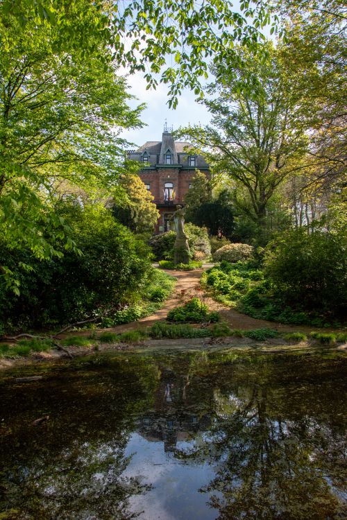 Free A picturesque brick house surrounded by lush greenery, reflected in a serene pond. Stock Photo
