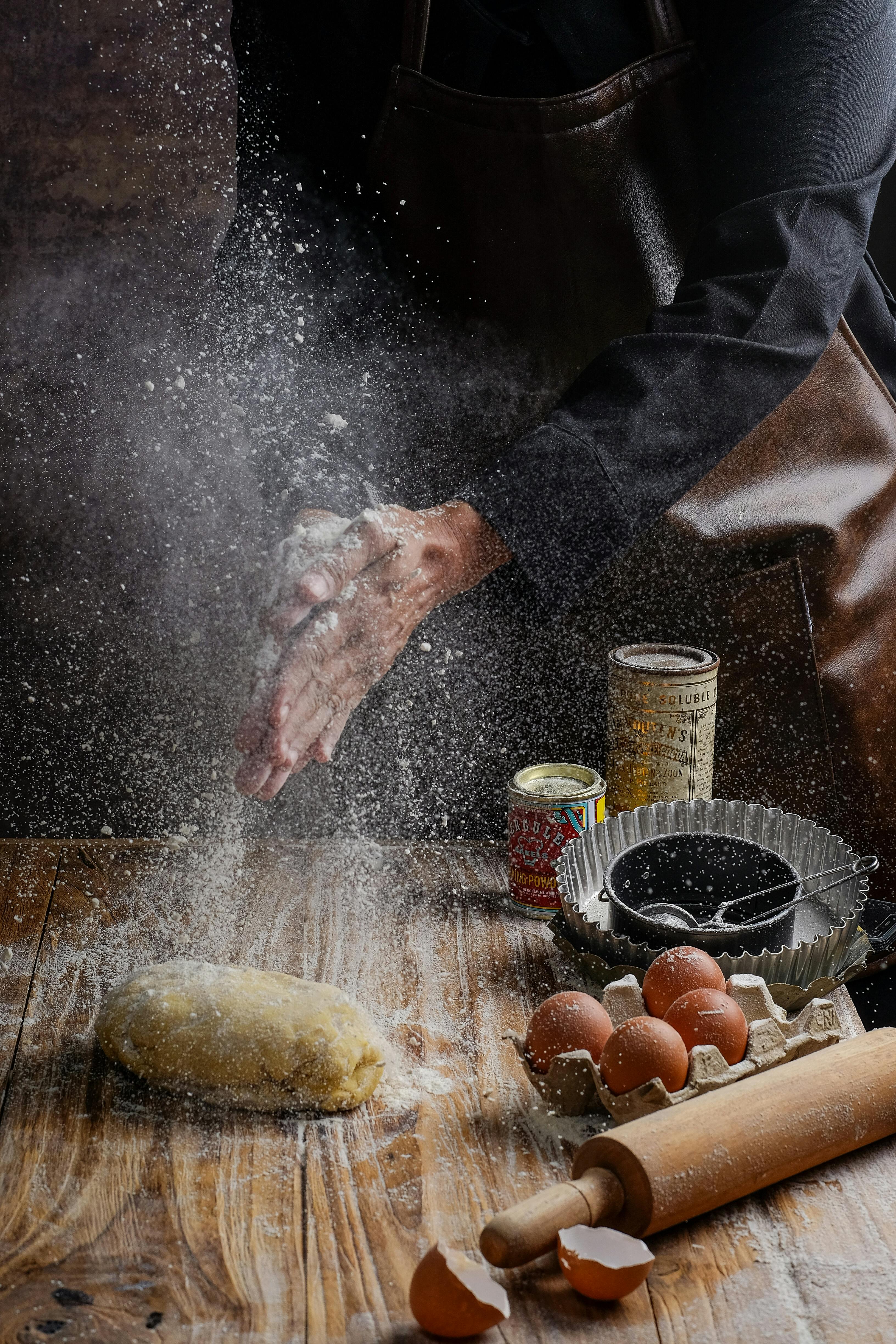 Hands clap flour over dough with baking ingredients on a rustic table.