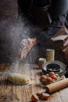 Hands clap flour over dough with baking ingredients on a rustic table.