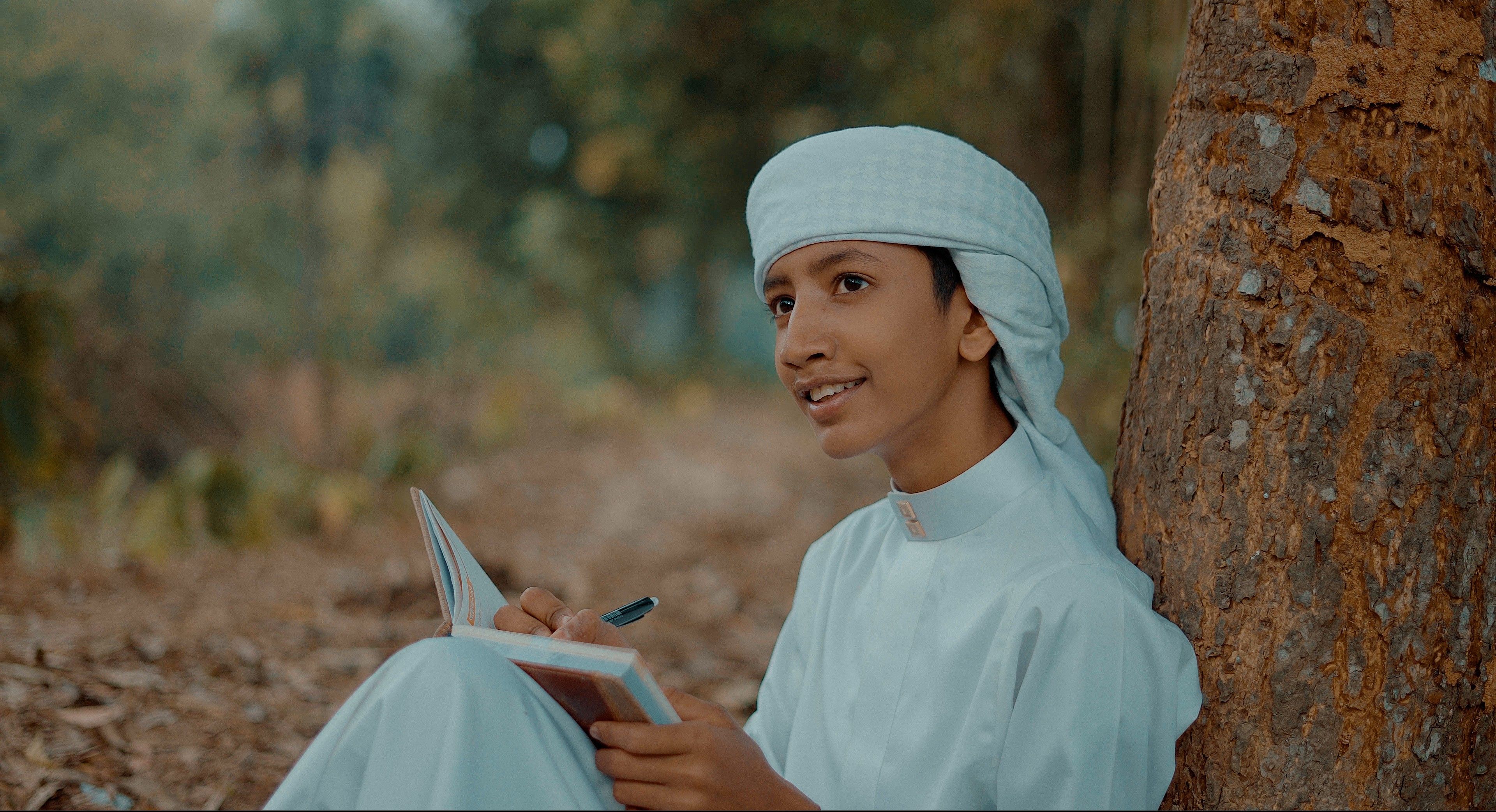 Middle Eastern boy in traditional attire writing in a notebook under a tree.