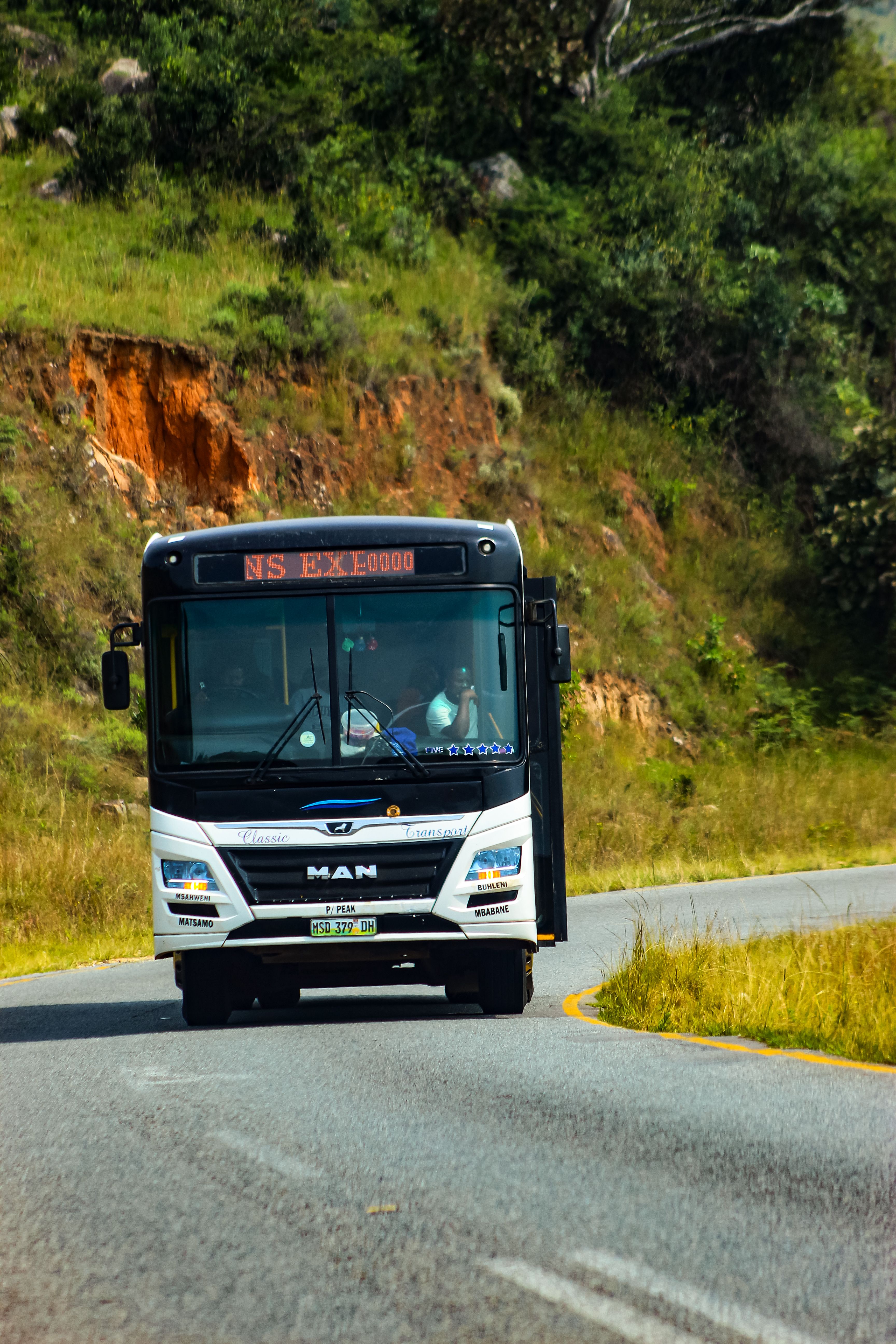 A bus traveling on a scenic road in Manzini, Eswatini, surrounded by lush greenery.
