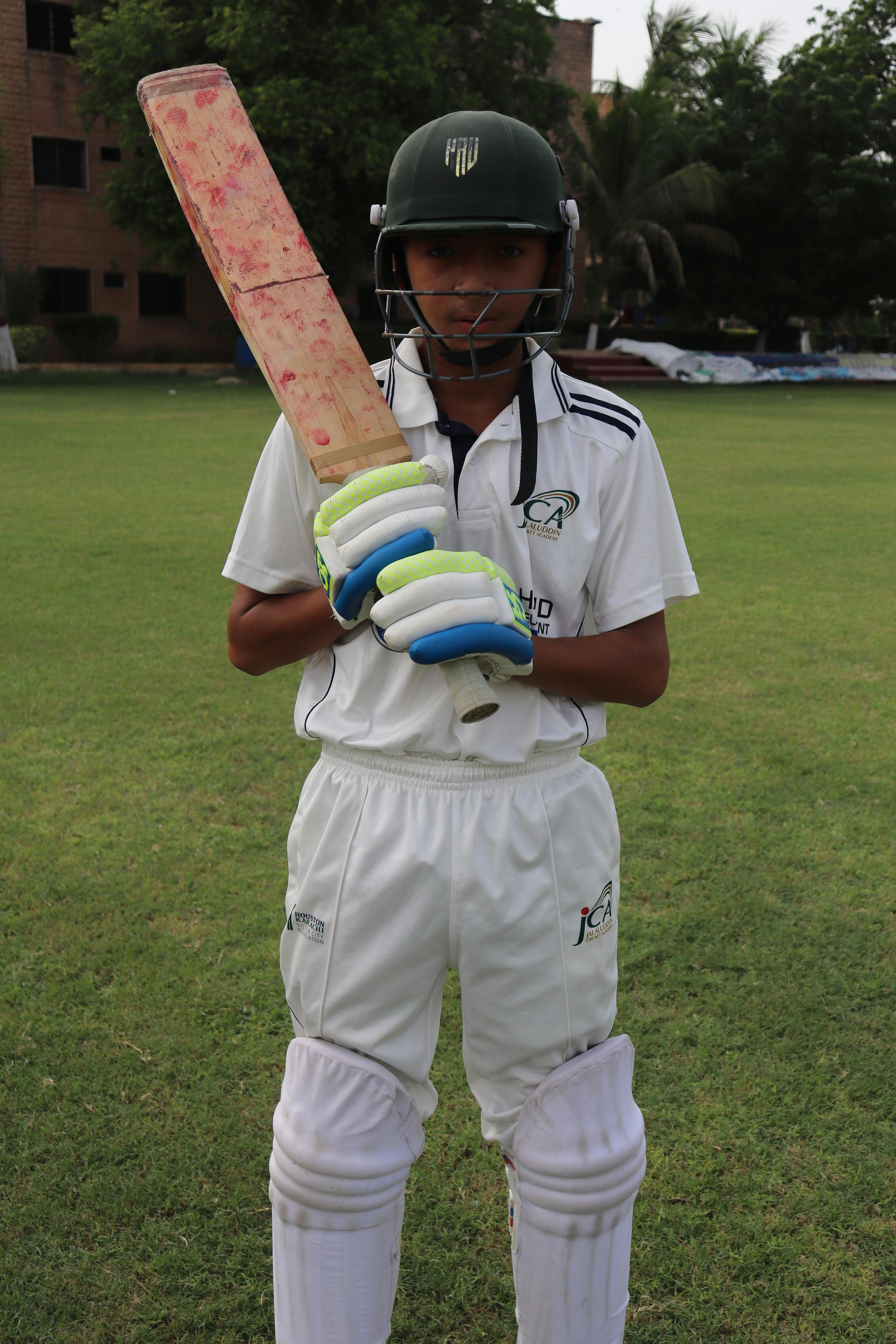 Teenage cricketer wearing full gear and holding a bat on a grassy field.