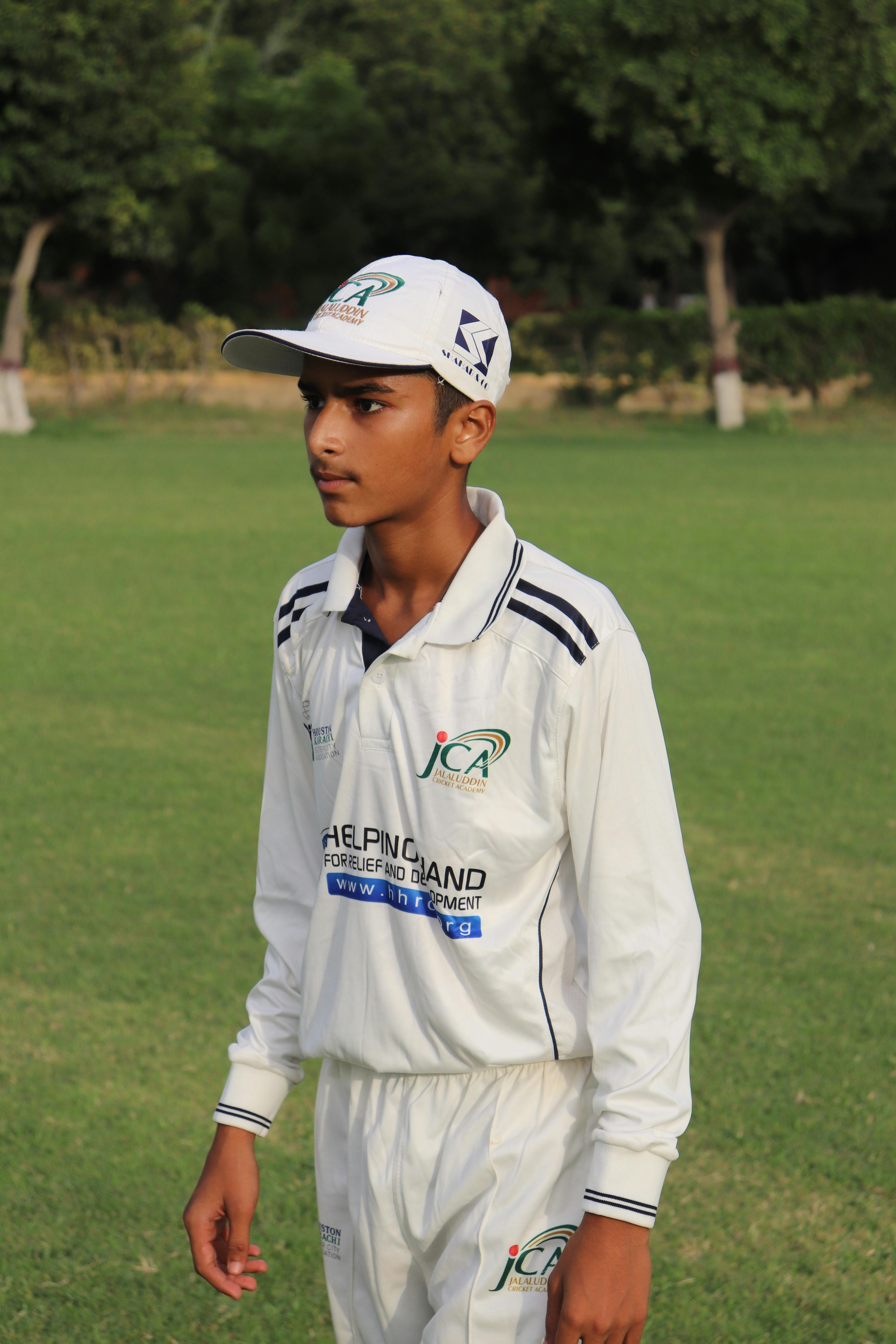 A teenage cricketer in uniform on a sunny day at a cricket ground.
