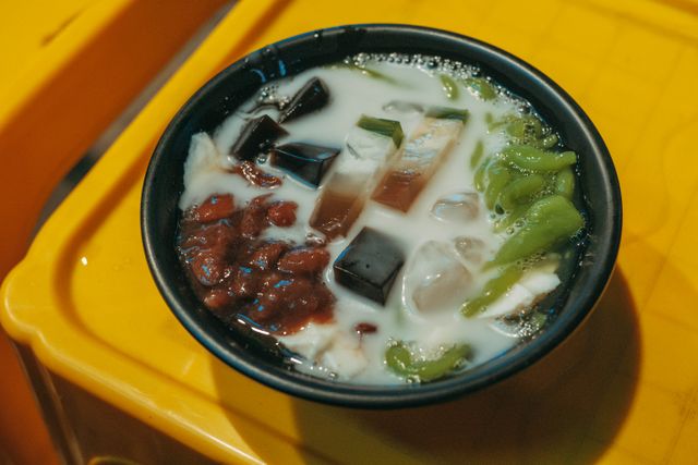 A delicious bowl of Asian dessert featuring colorful jelly cubes, red beans, and creamy milk on a vibrant yellow background.