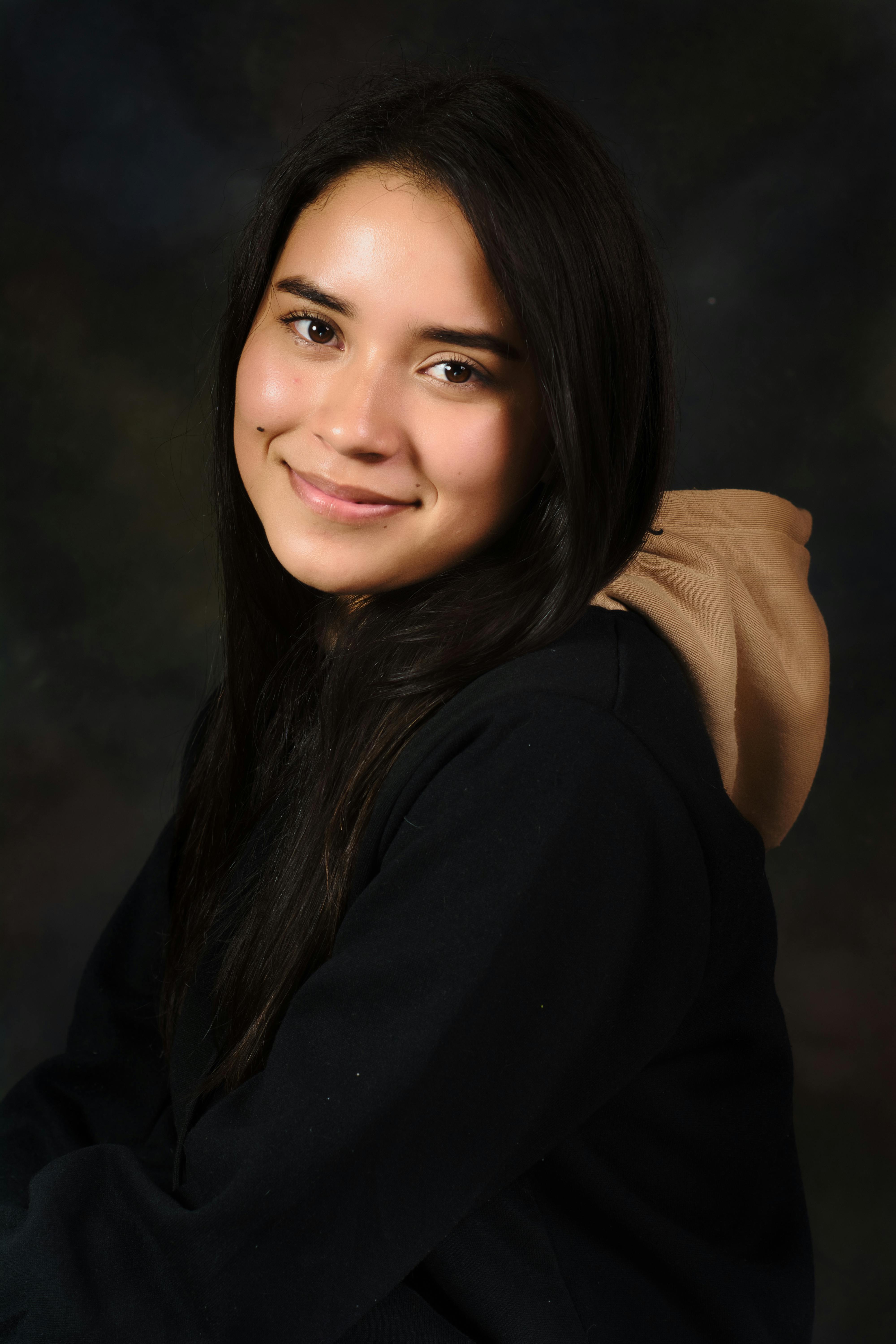 A young woman with long dark hair smiling, wearing a black hoodie, photographed indoors.