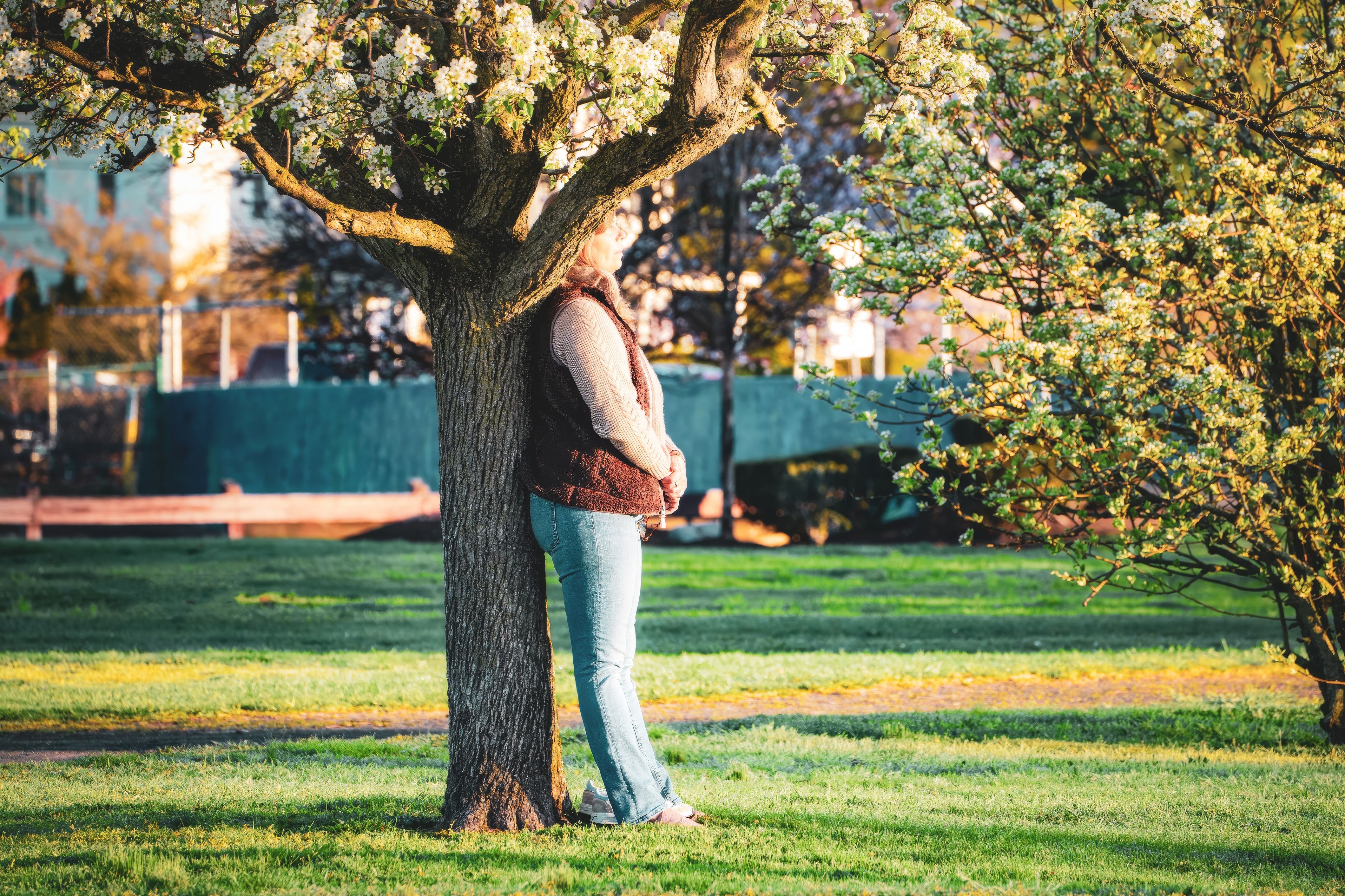 A woman leans against a flowering tree in a sunny park, enjoying a peaceful spring day outdoors.