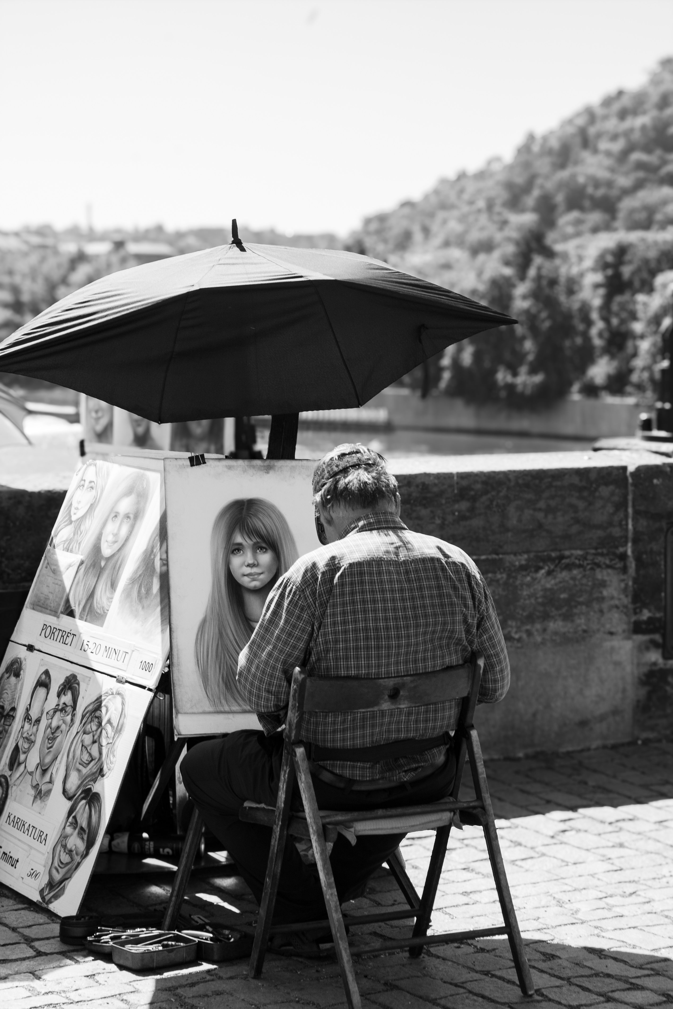 A street artist sketching a portrait under an umbrella by the riverside in a picturesque setting.
