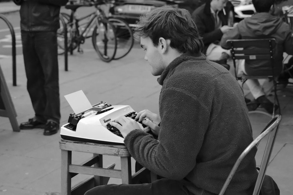 A man typing on a typewriter outdoors in a bustling street scene in England.