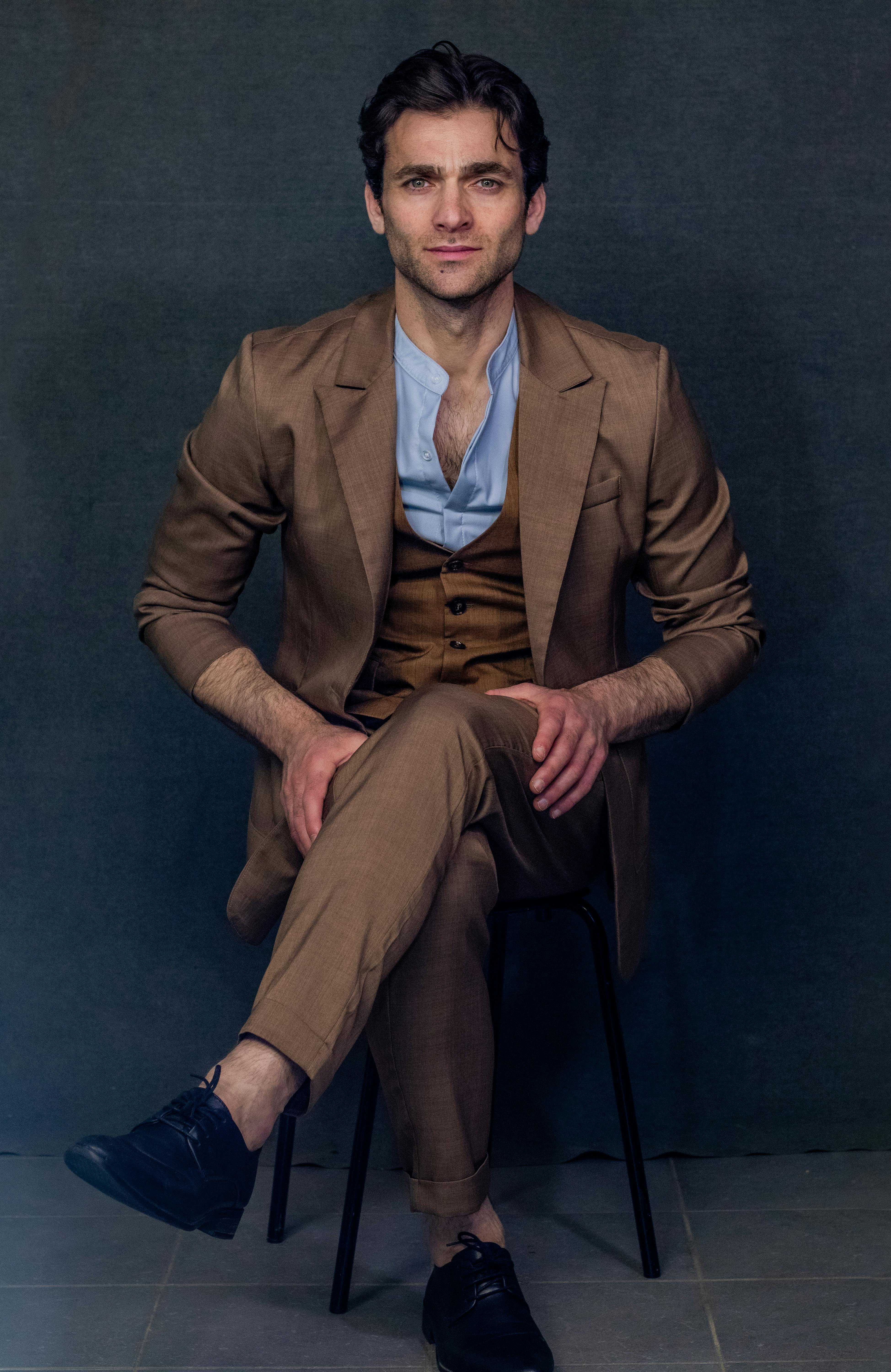 Confident man posing in a brown suit with blue shirt against a dark background.