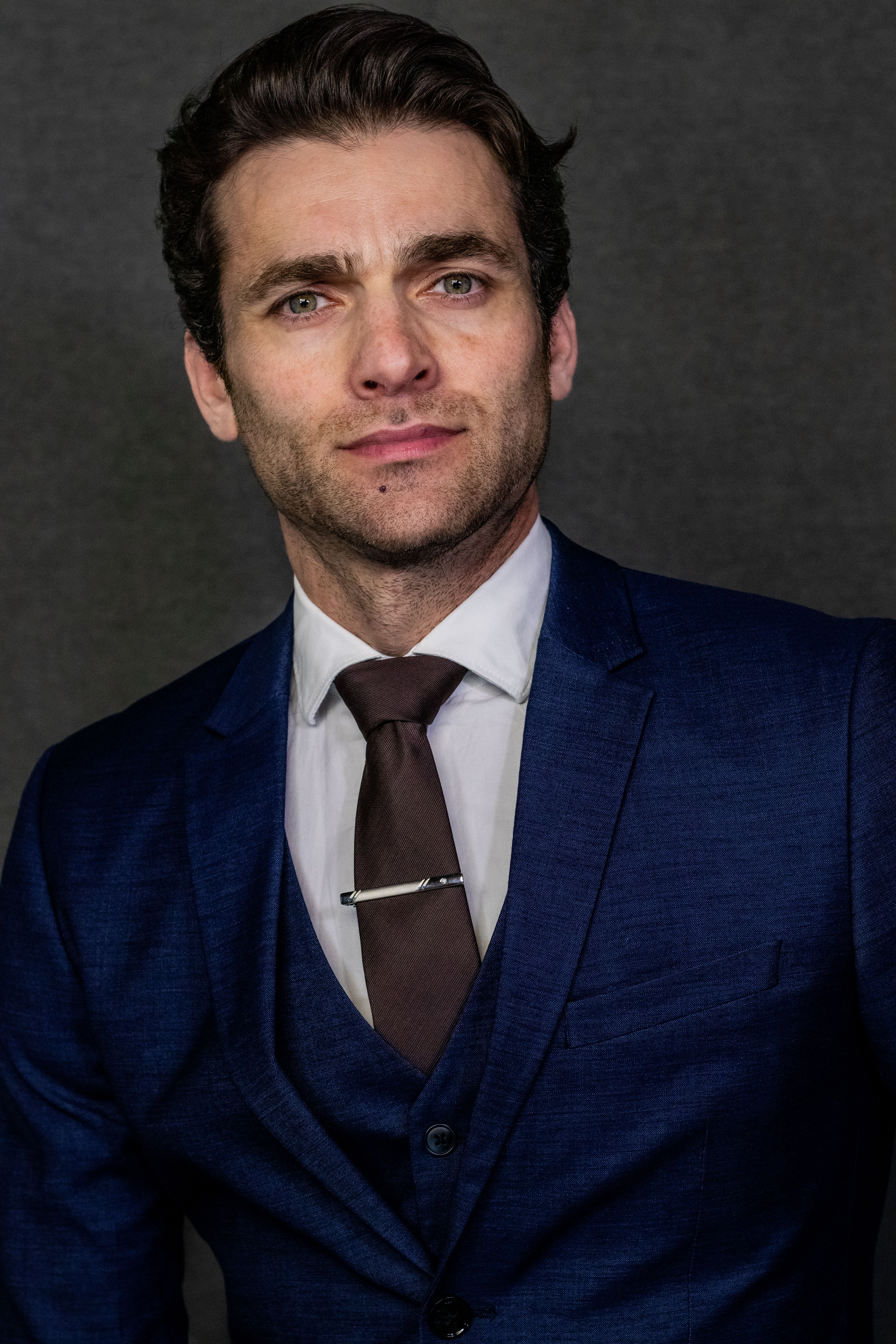Professional portrait of an adult man in a blue suit and tie against a dark background.