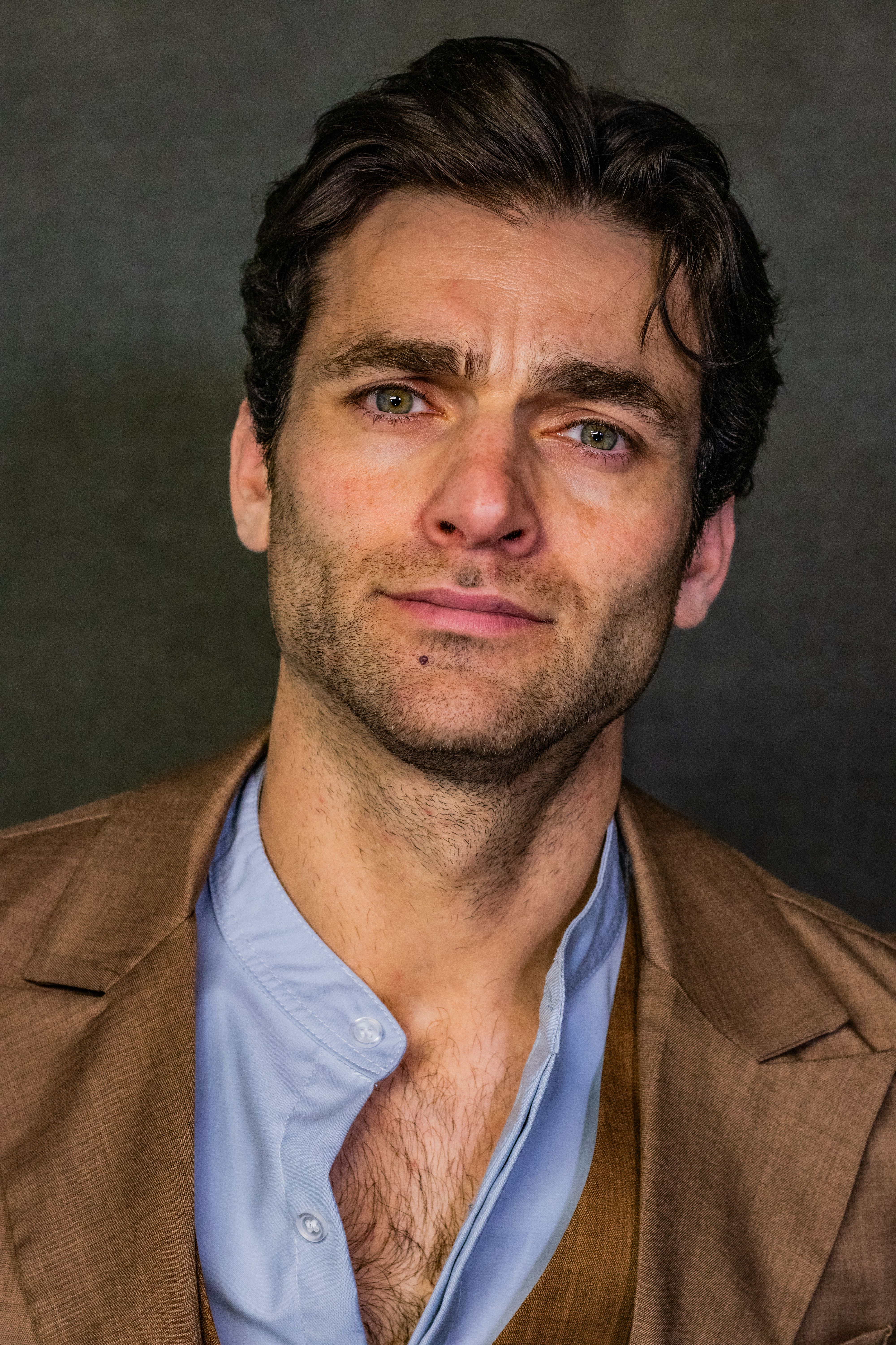 A close-up portrait of a thoughtful adult man wearing a brown jacket.