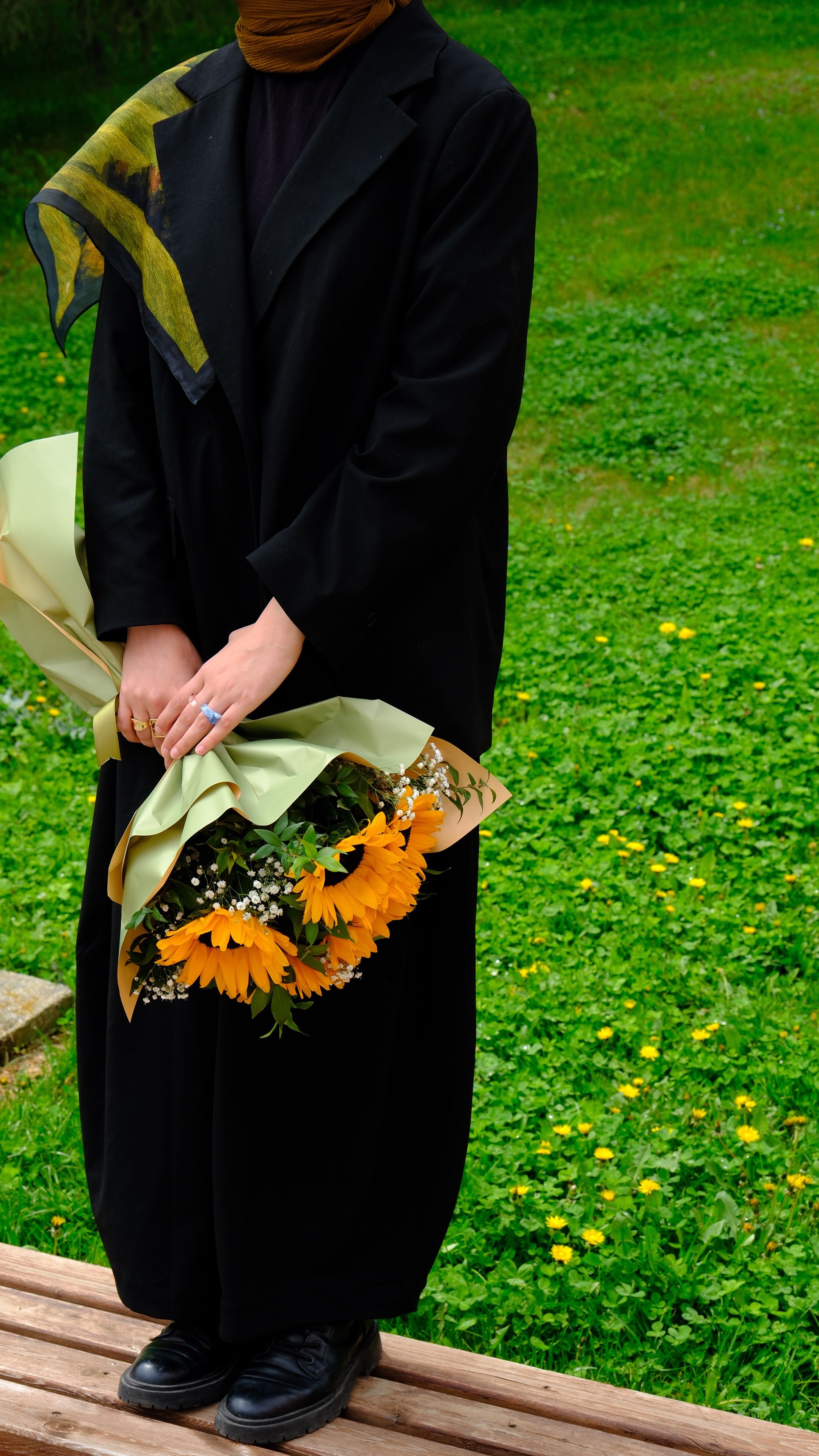 A woman in a black hijab holds a sunflower bouquet on a grassy field.