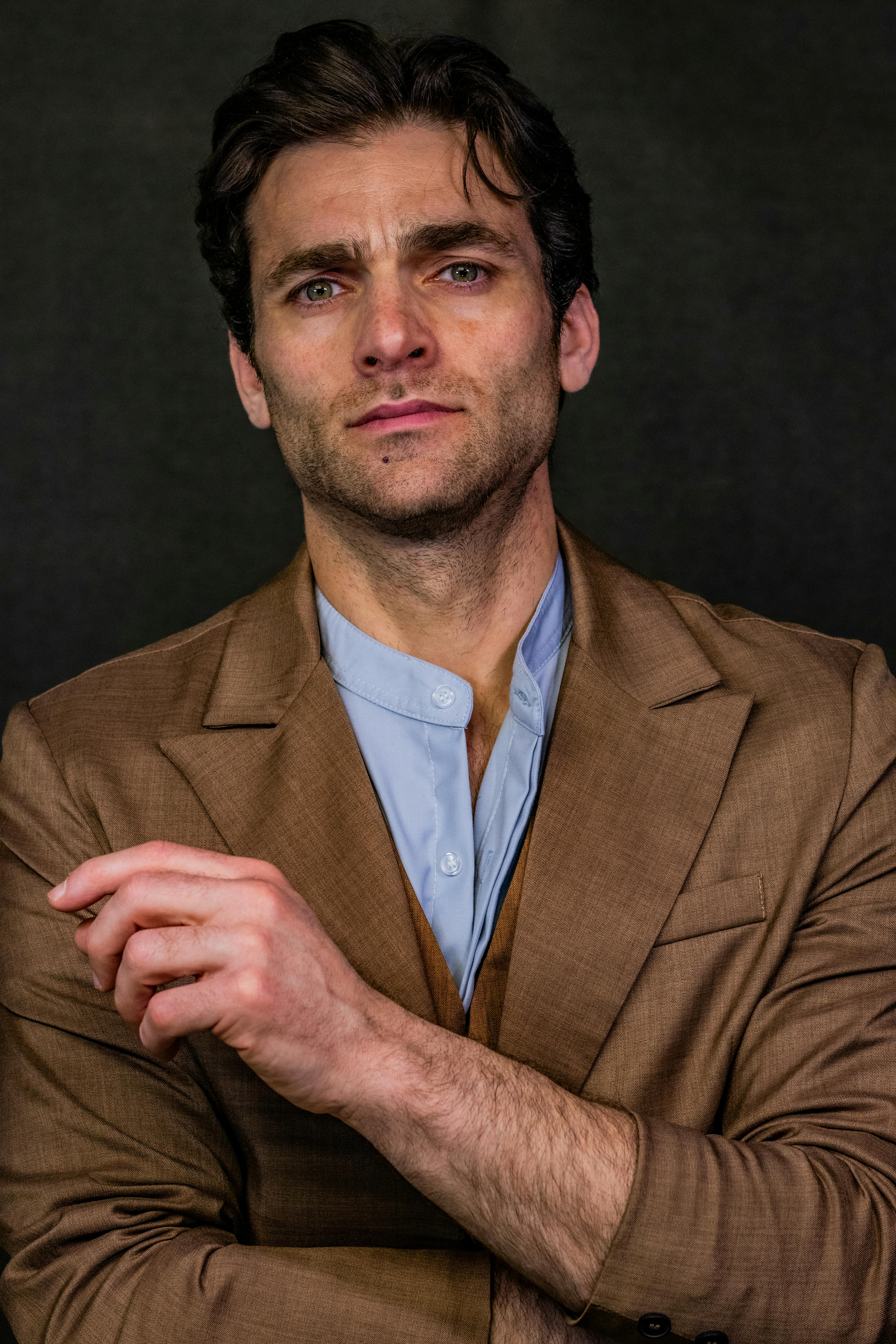 Close-up portrait of a man in a brown suit against a dark backdrop, exuding confidence.