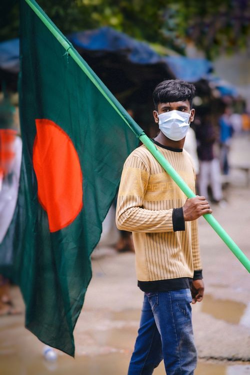 Free A young man wearing a mask holds the Bangladesh flag in an urban outdoor setting, reflecting national pride. Stock Photo