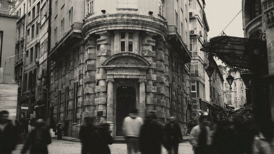 Vista histórica de una calle urbana con un edificio de arquitectura antigua; los transeúntes aparecen borrosos.