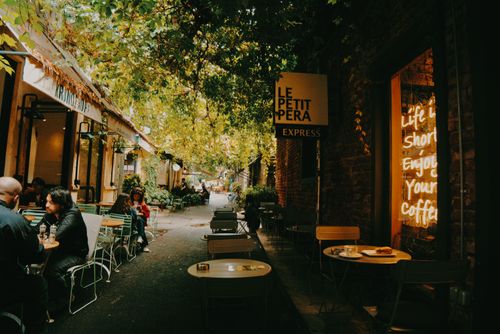Free A cozy outdoor café scene with people enjoying coffee under trees in a charming alleyway. Stock Photo