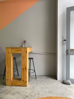 A minimalist apartment corner with a wooden table, stools, and a vase on concrete floor.