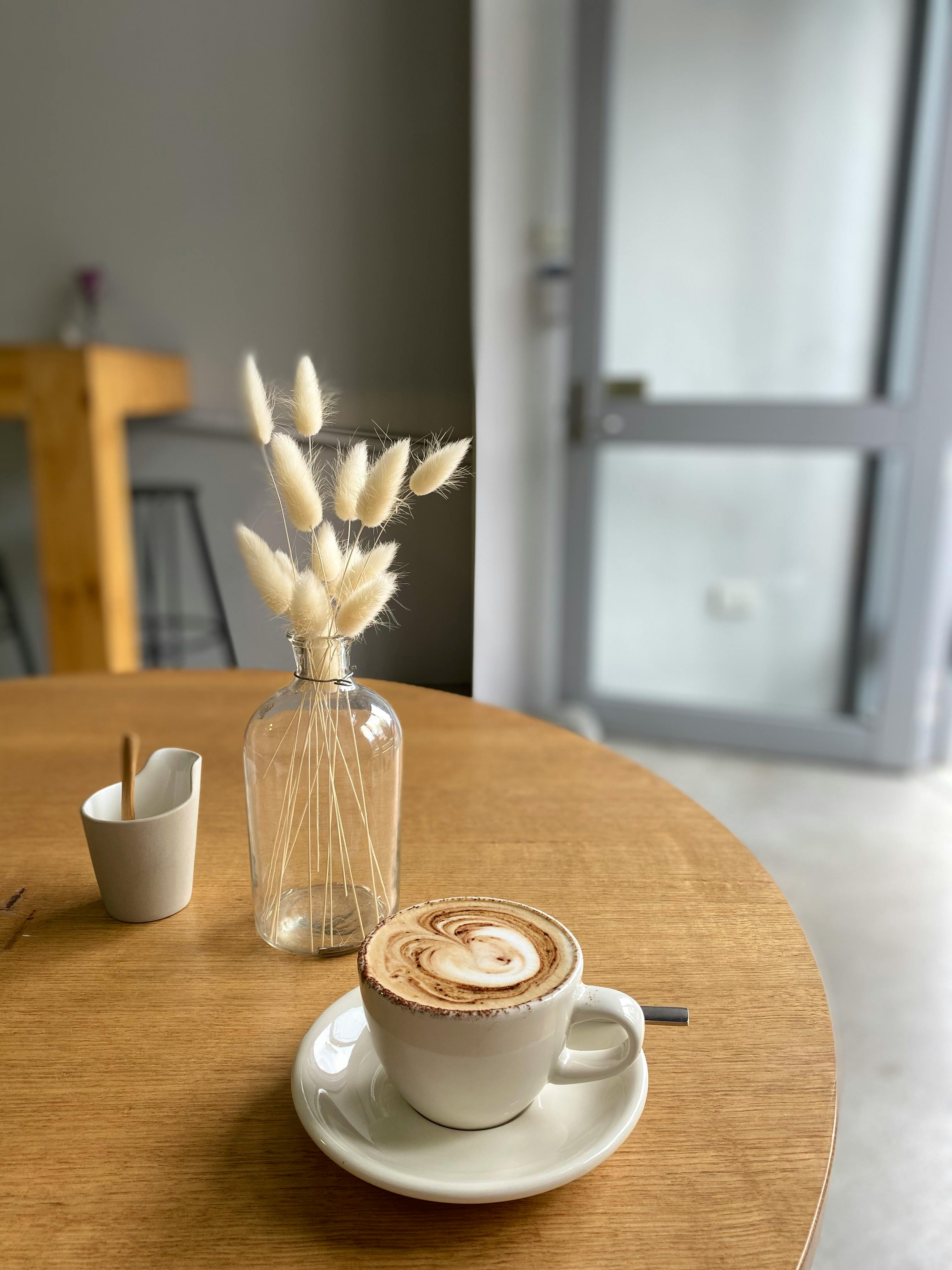 white ceramic cup on saucer beside flower vase on table