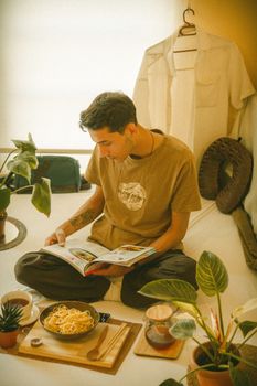 Young man sits reading on a bed, surrounded by plants and a meal, creating a cozy indoor atmosphere.