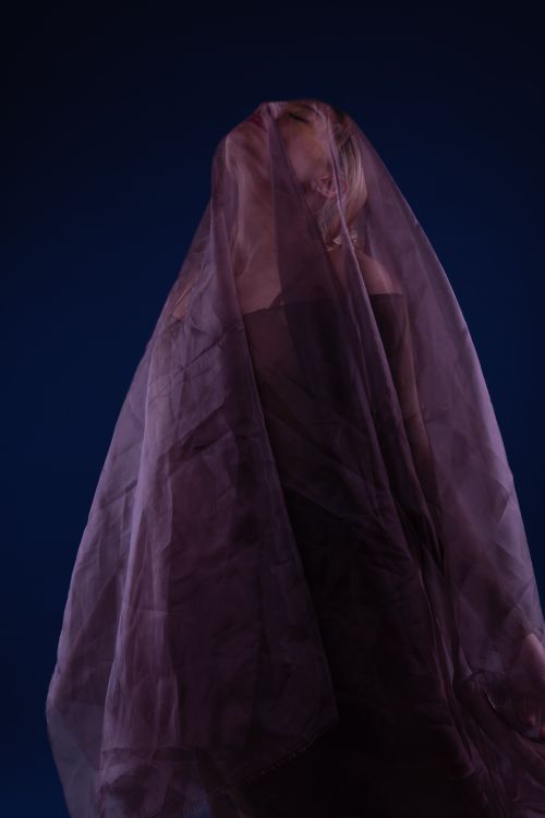 Free A dramatic artistic portrait of a woman enveloped in a translucent purple veil against a dark blue backdrop. Stock Photo