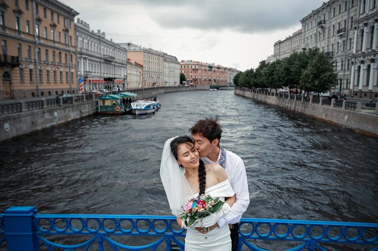 Couple Standing On Bridge Wth Blue And White Boat On River