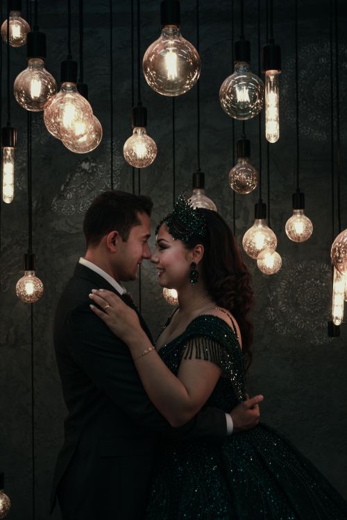 Free A couple in elegant attire embraces romantically under decorative hanging light bulbs. Stock Photo