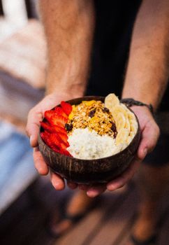 A vibrant breakfast bowl featuring strawberries, bananas, granola in a coconut bowl, held outdoors.