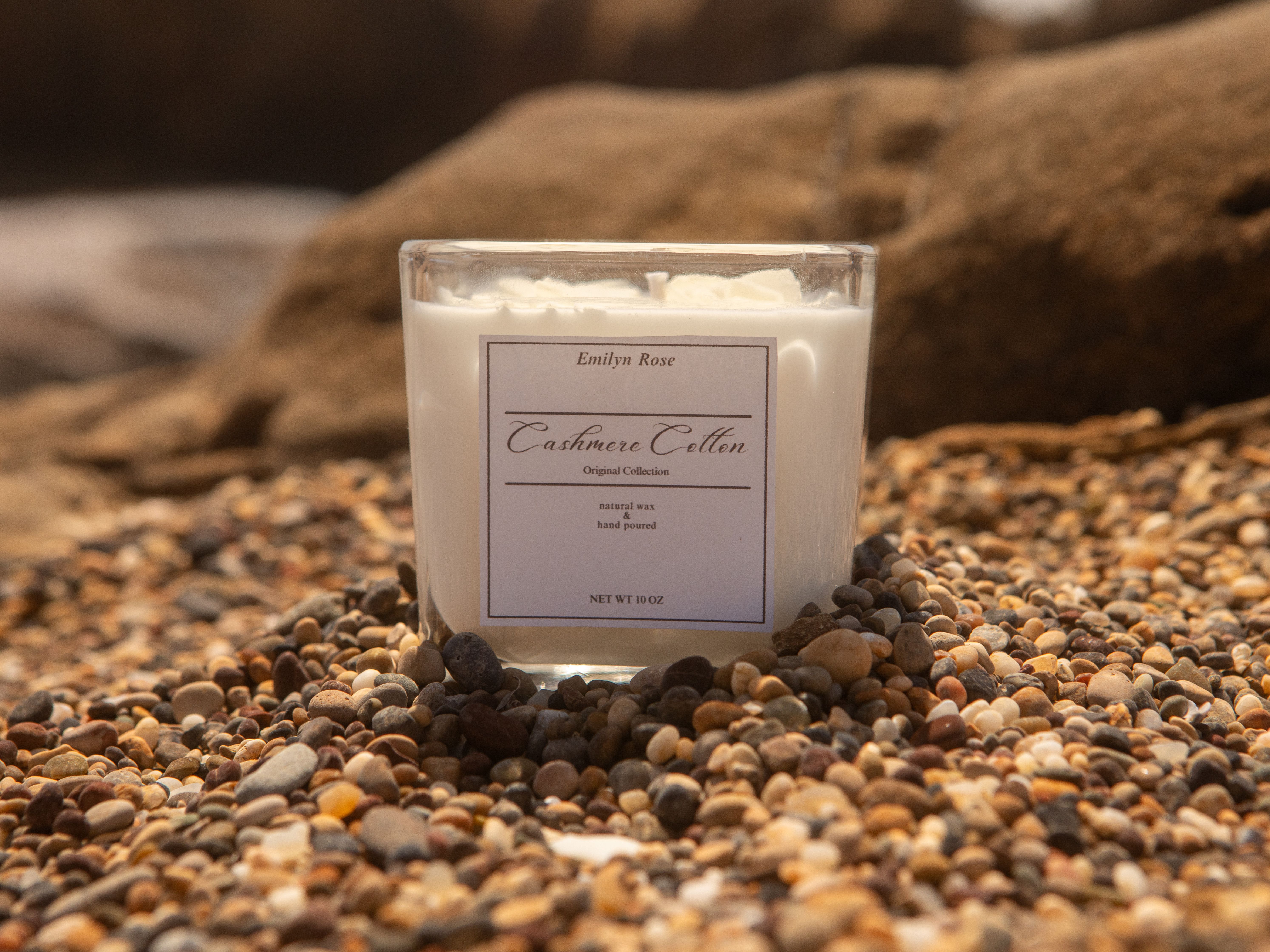 A cashmere cotton scented candle placed on beach pebbles, evoking a tranquil coastal vibe.