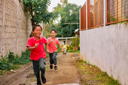 Free Two smiling children running joyfully on a sunny day through a rustic alleyway. Stock Photo
