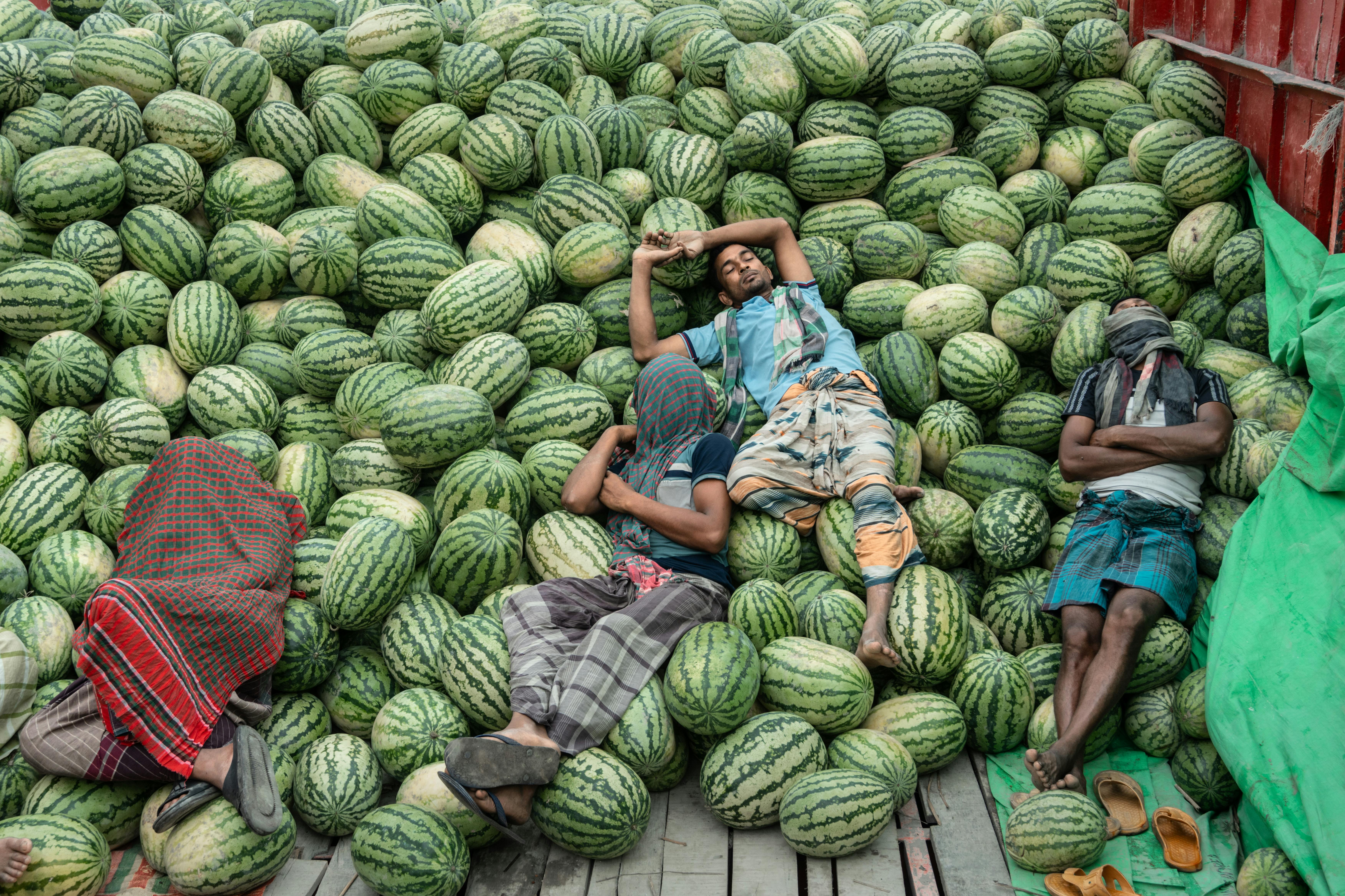 Men resting on a pile of watermelons in a vibrant outdoor market scene, conveying summer vibes.