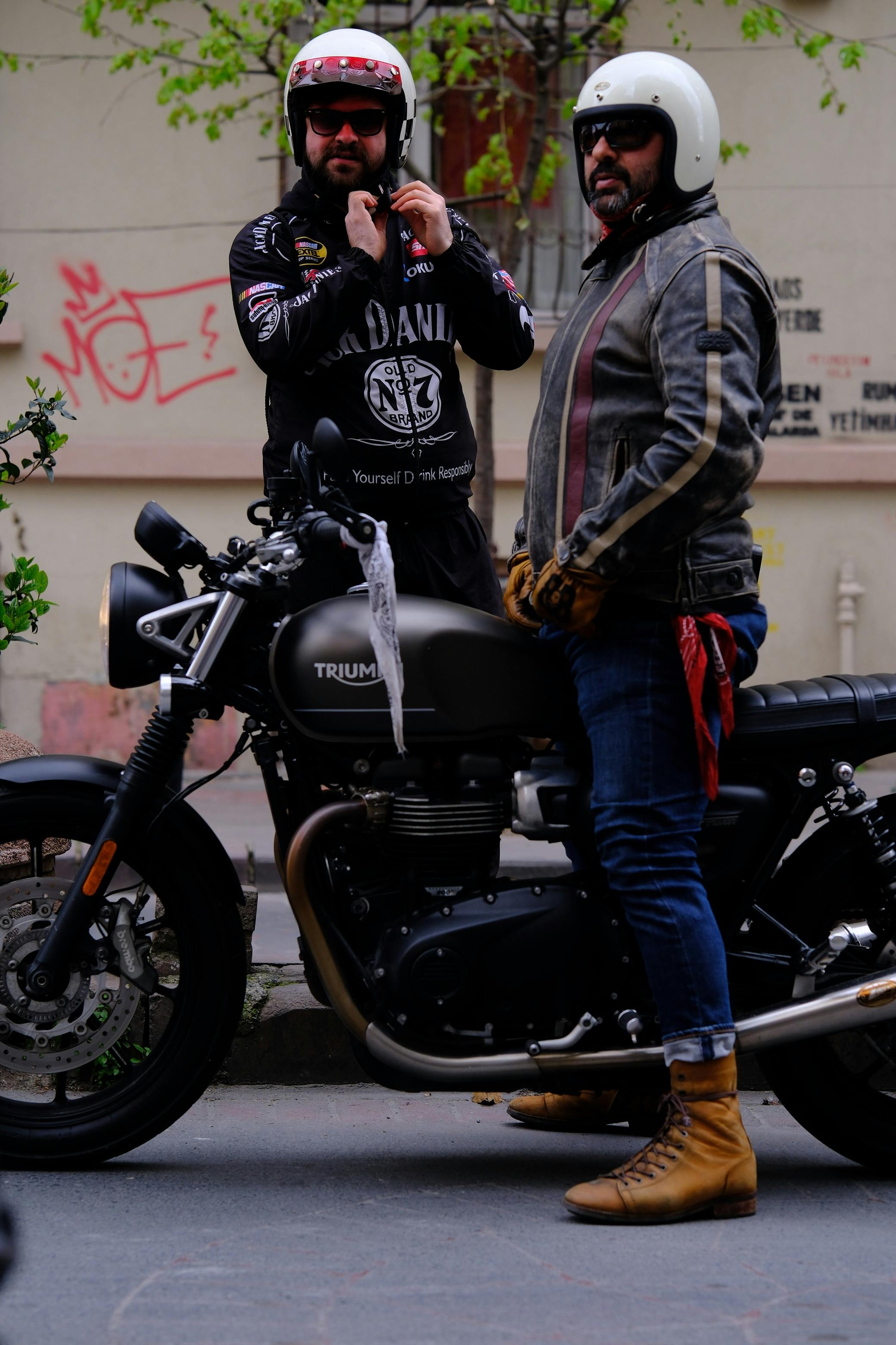 Two men wearing helmets and jackets stand beside a Triumph motorcycle on a city street.