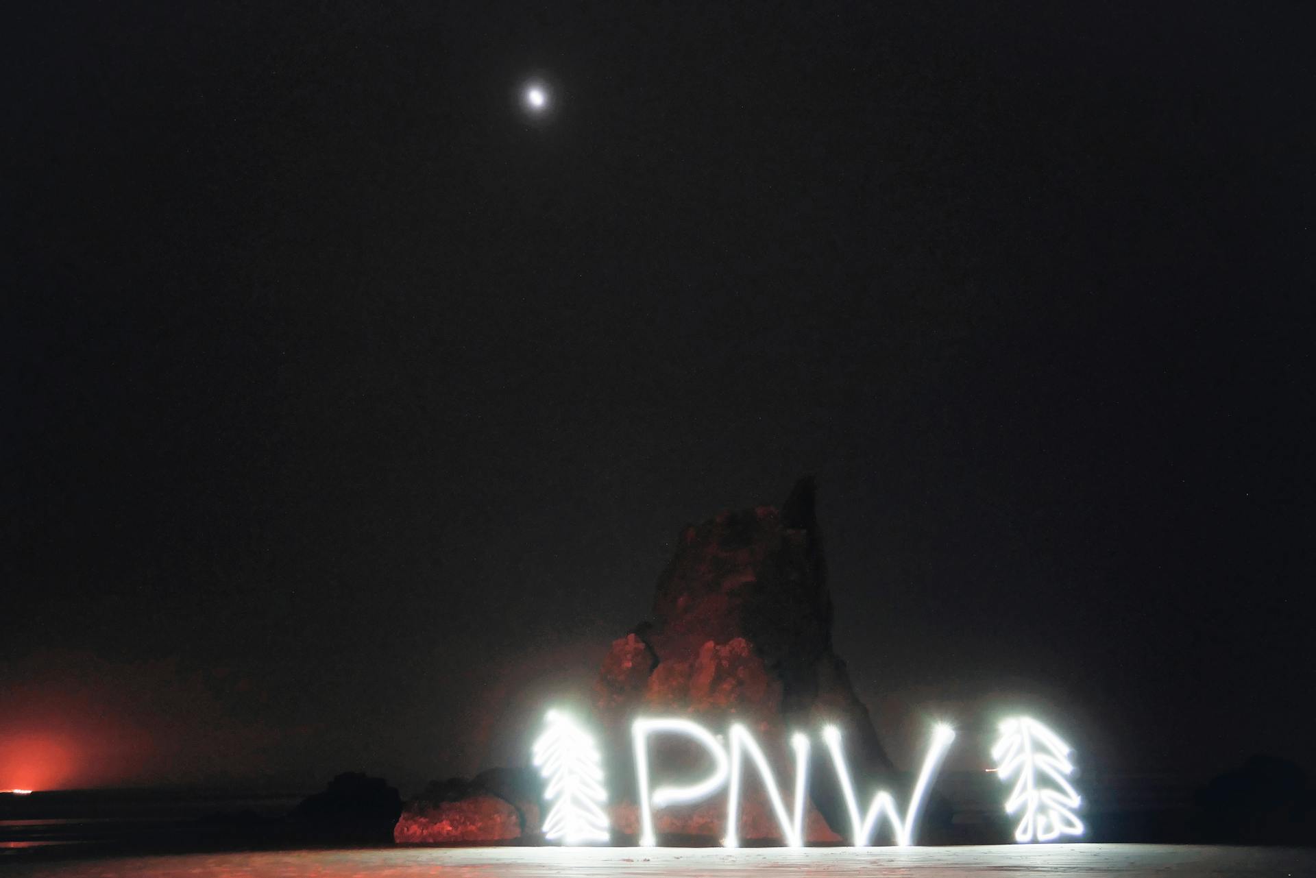 Abbreviation PNW between fir shaped decorations illuminating with white neon lights and located against rocks at night under dark sky and bright moon Pacific Northwest traveling