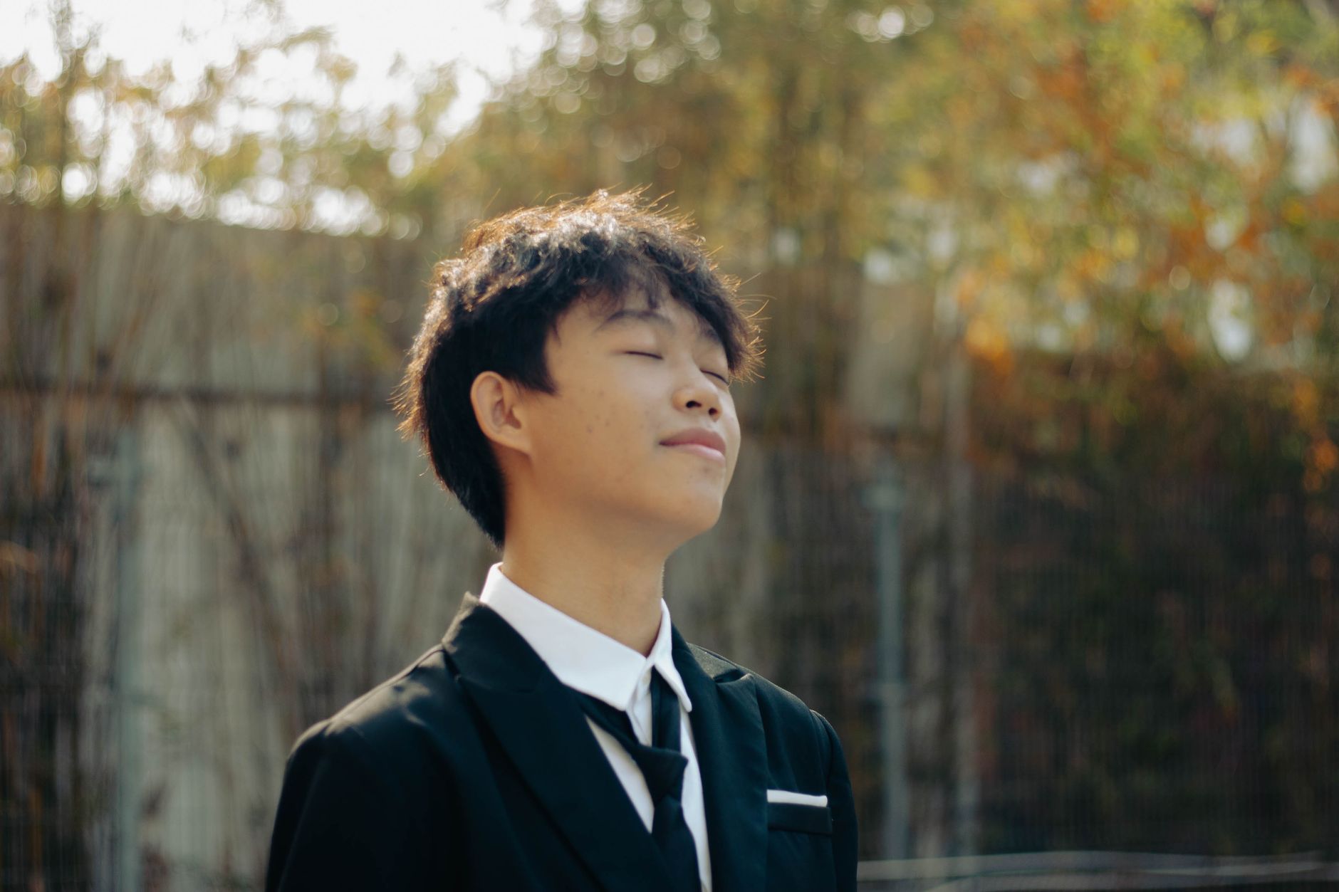 A young man in a suit enjoys the sunlight with a peaceful expression outdoors.