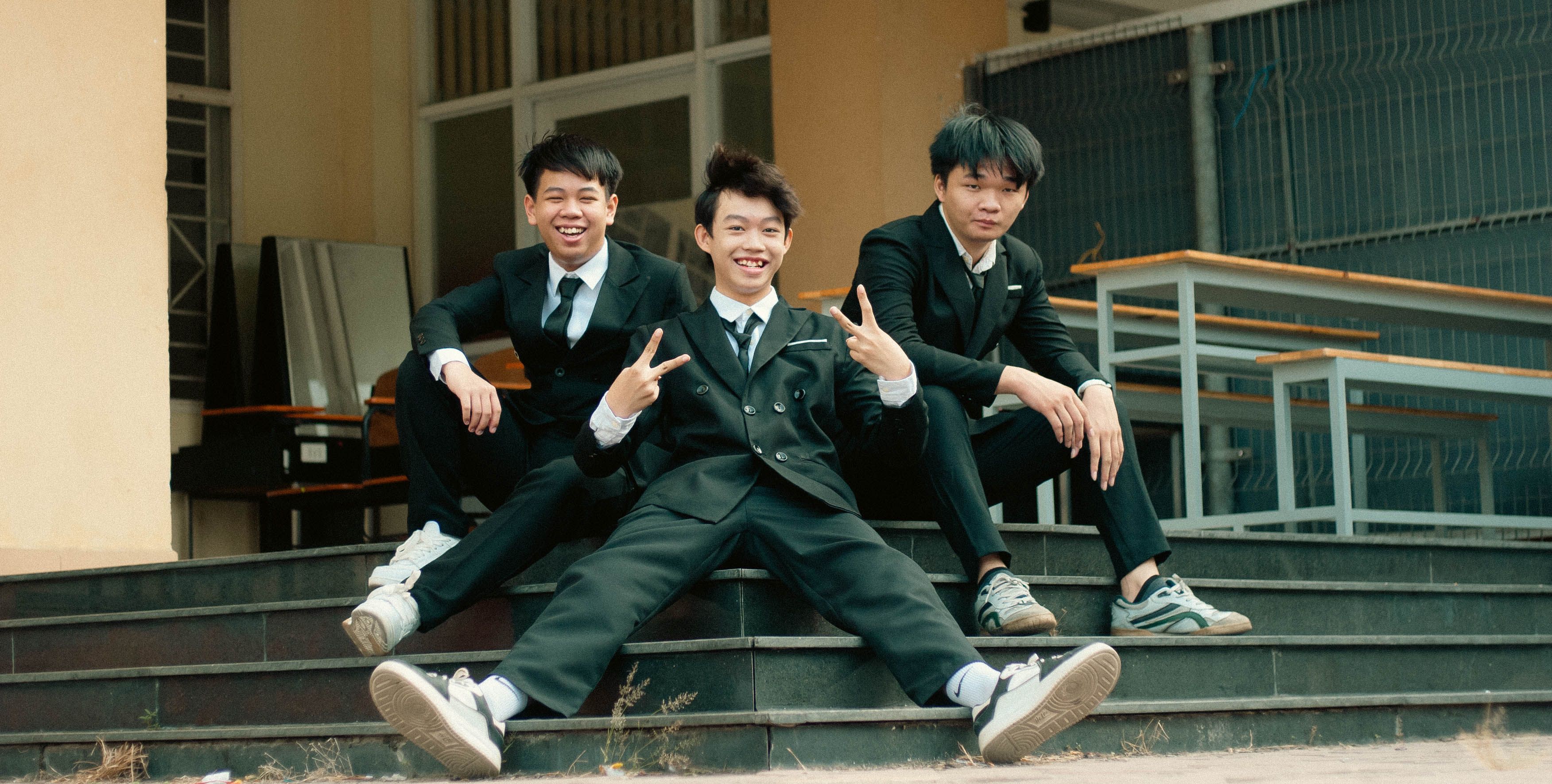 スーツを着た3人の若い男性が、屋外の階段で笑顔でポーズをとっている。